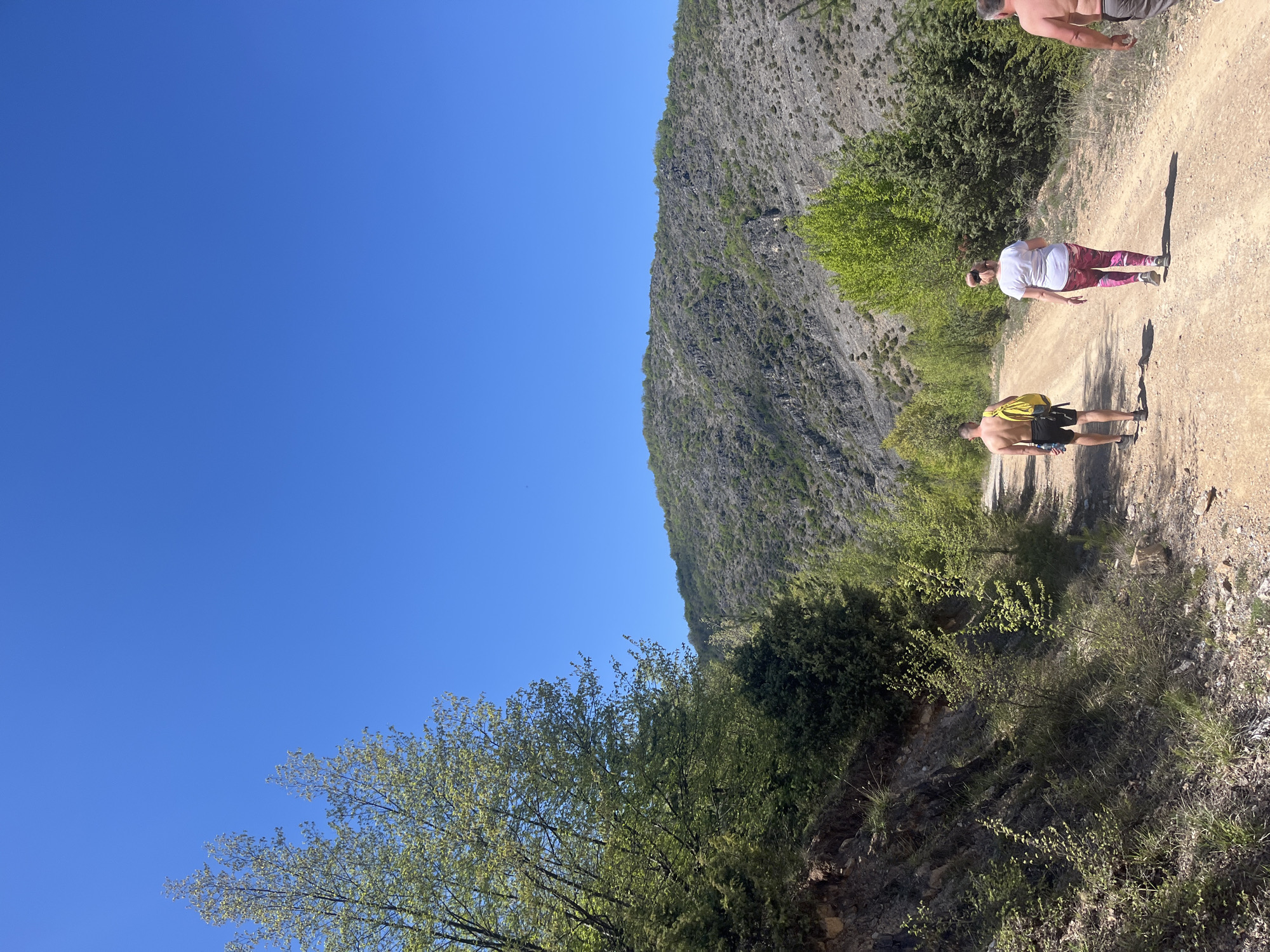 The image shows a dirt path in a natural environment, possibly a park or wilderness area in Polska Skakavitsa, Bulgaria. A rocky hill with sparse vegetation rises on the left side of the path, and trees/shrubs line the path on the right. There are three people on the path, two with no shirts. One man is walking. A woman is walking. The weather appears to be clear and sunny, as evidenced by the bright blue sky.