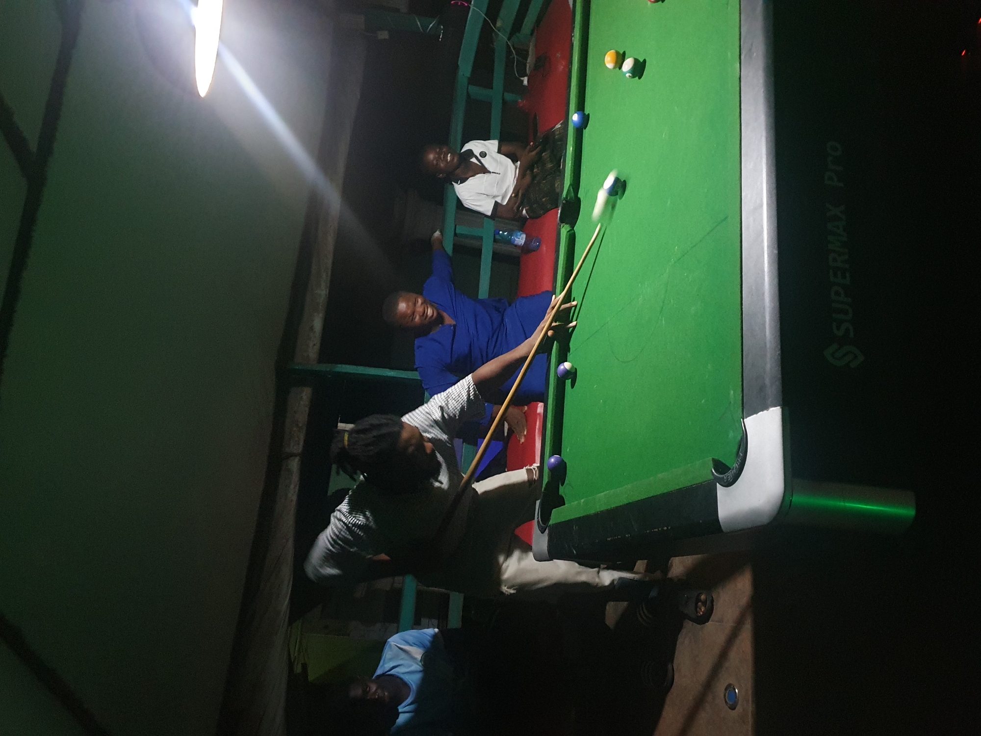 In Lokoja, Nigeria, a group of individuals are engaged in a game of billiards. The scene unfolds at night, illuminated by a ceiling light that casts a warm glow on the green billiard table. Four people are visible, with one woman in the foreground actively playing, poised to strike a ball with her cue. She wears a striped shirt and light-colored pants. To her left, a man in a blue shirt watches the game. Behind the player, two individuals are seated and appear to be laughing or cheering. One is dressed in a blue outfit, and the other in a white polo shirt. The billiard table is set with several colorful balls scattered across its surface, and the overall environment suggests a casual, recreational gathering.