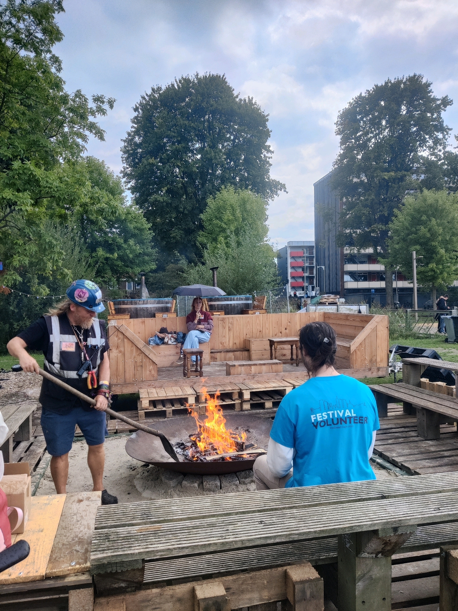 The image depicts an outdoor gathering in the city of Tilburg, Netherlands, possibly at a community event or festival, given the "FESTIVAL VOLUNTEER" text on a blue t-shirt. In the foreground, a person wearing a hard hat and a high-visibility vest is tending to a bonfire with a shovel. They have long hair and a beard, and are dressed in shorts and a black t-shirt. The bonfire is in a large metal pit, surrounded by wooden seating and structures made of pallets. In the midground, another person, seen from behind, is wearing the blue "FESTIVAL VOLUNTEER" t-shirt and light-colored pants, sitting and observing the fire. Further back, a person is seated on a wooden bench under a black umbrella, holding a book or tablet. Behind them, wooden structures, possibly part of an outdoor stage or seating area, are visible, along with some trees and buildings in the background. The sky is overcast, suggesting a cloudy day. The environment appears to be a recreational or communal space, with a mix of natural elements like trees and man-made structures for social interaction.