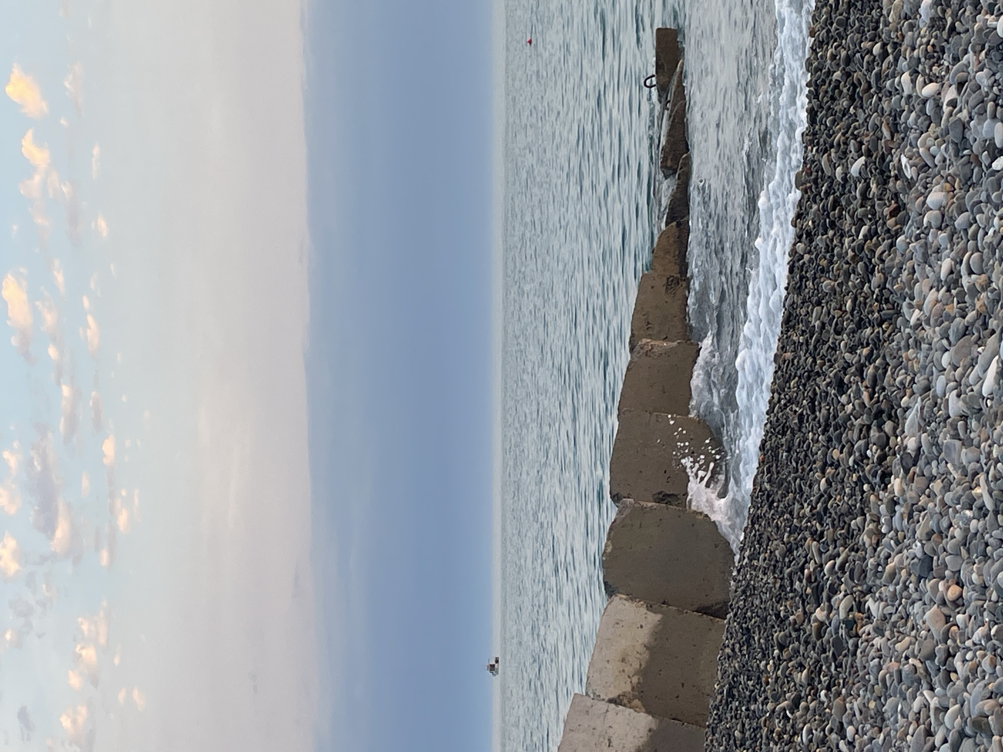 The image depicts a serene coastal scene at dusk. In the foreground, a pebble beach stretches across the bottom of the frame. A breakwater made of concrete blocks juts out into the calm sea, with gentle waves lapping at its base. In the distance, a small boat is visible on the horizon, and a red buoy floats in the water to the right. The sky is a soft blend of blue and pale pink, with scattered clouds, indicating the fading light of evening. The overall atmosphere is peaceful and tranquil.
