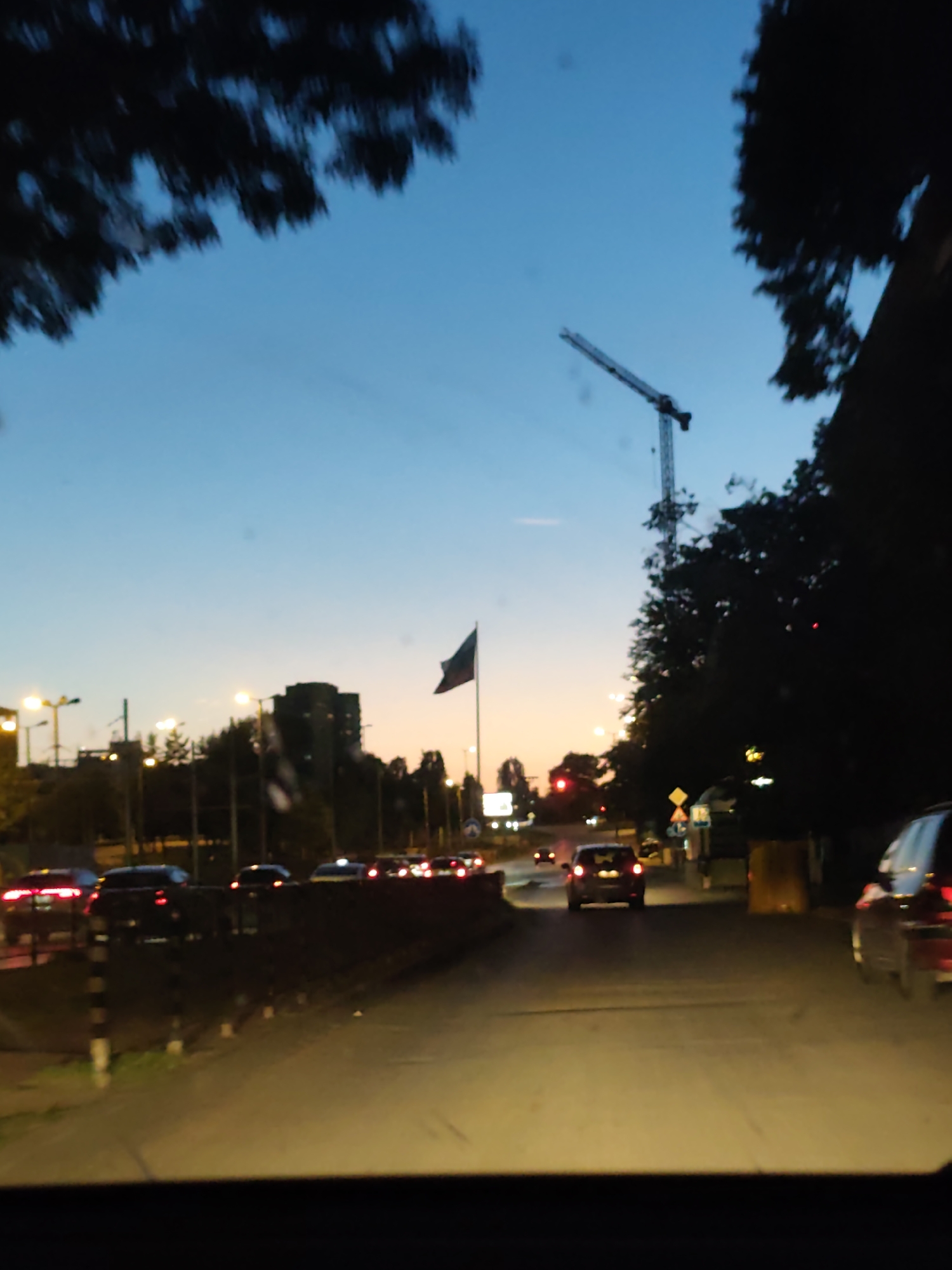 The image depicts a street scene at dusk in Sofia, Bulgaria. The view is from inside a vehicle, looking out through the windshield. The sky is a gradient of blue to orange. A Bulgarian flag is visible in the middle distance, alongside buildings, streetlights, and a construction crane. The road is illuminated by car headlights and streetlights, showing several cars in traffic. Trees frame the top of the image. Street signs are present on the right side of the road.