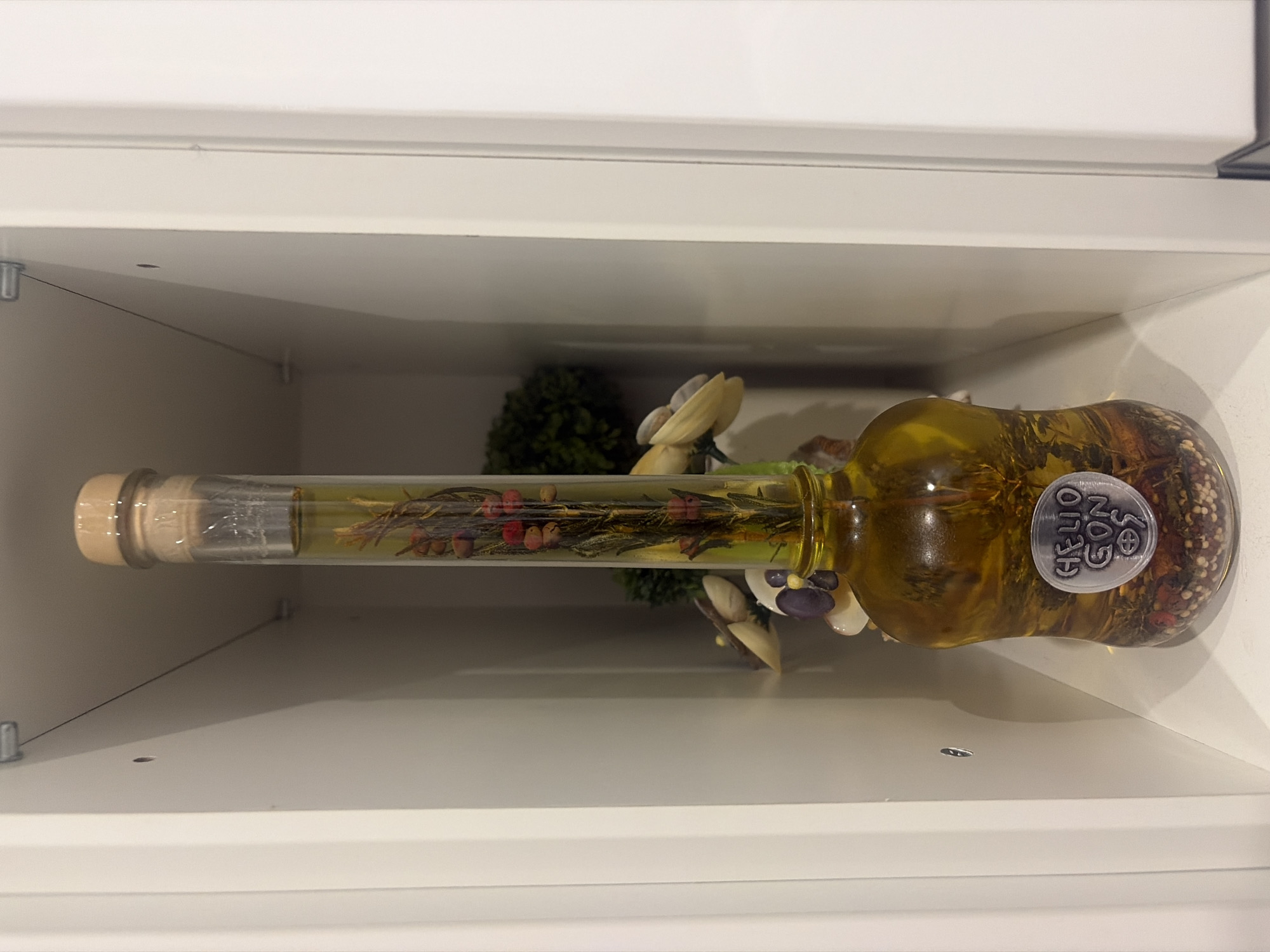 A narrow, clear glass bottle with a cork stopper, filled with a yellow liquid and suspended botanicals, stands upright on a white shelf within a white recessed cabinet. The botanicals appear to be a sprig of rosemary with small red berries, and a variety of dried spices or seeds at the bottom of the bottle. A circular, metallic-looking label with the embossed text "HELIO GON 9" is affixed to the front of the bottle. Behind the bottle, a blurred green decorative bush and some seashell-like objects are visible. The scene is lit from the front, casting a subtle shadow on the left side of the cabinet. The location is identified as Podgumer, Bulgaria.