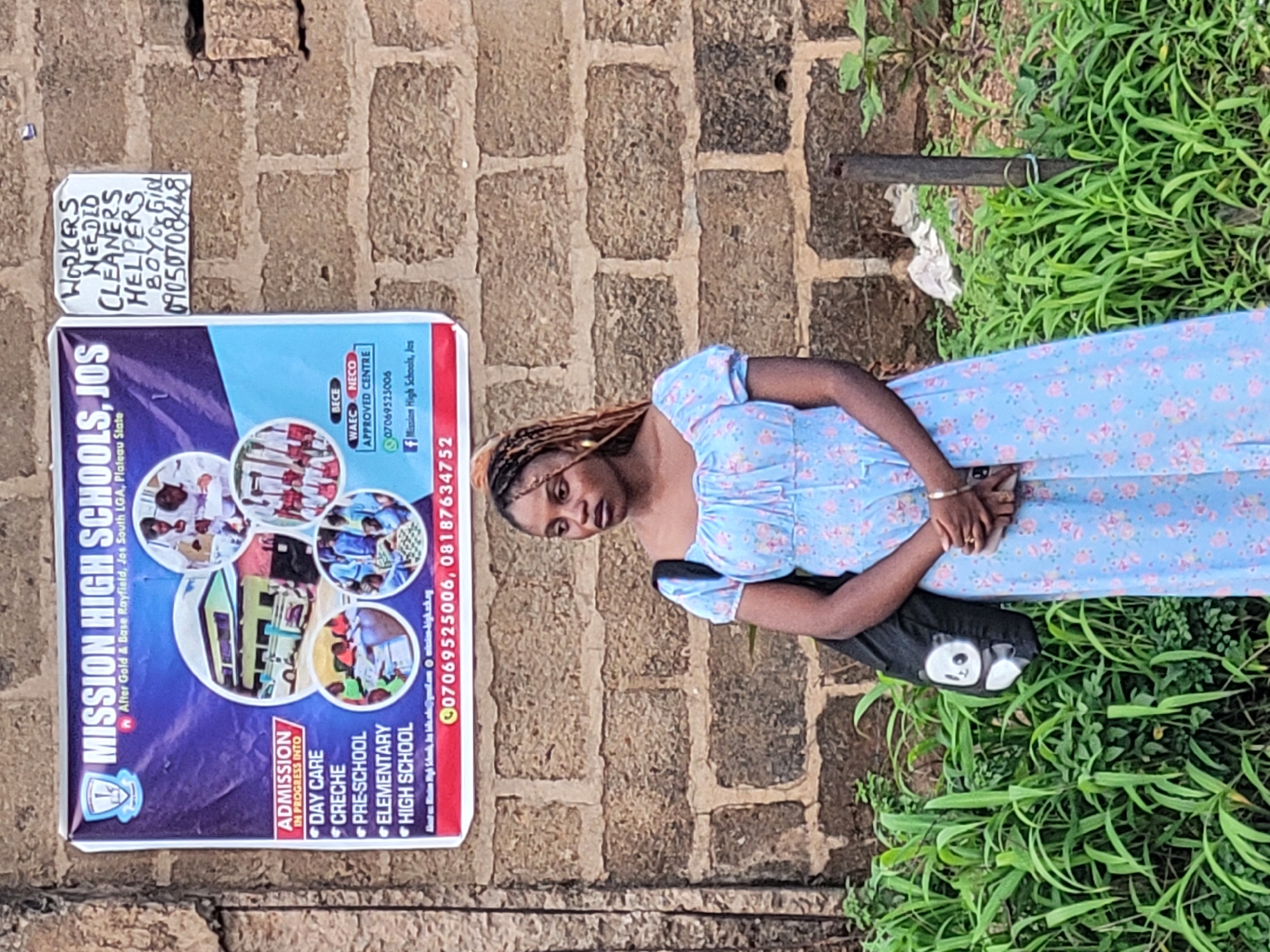 The image shows a young woman with braided hair wearing a light blue dress with a floral pattern. She is standing outside, leaning against a brick wall, with lush green grass in the foreground. Her hands are clasped in front of her, and her gaze is directed downwards, suggesting a thoughtful or perhaps somber mood.
Affixed to the brick wall is a colorful poster advertising "MISSION HIGH SCHOOLS, JOS". The poster lists the school's location as "After Gold & Base Rayfield, Jos South LGA, Plateau State" and announces "ADMISSION IN PROGRESS INTO" various levels, including Day Care, Creche, Pre-School, Elementary, and High School. It also provides contact numbers and mentions that the school is an "APPROVED CENTRE" for BECE, WAEC, and NECO examinations. Another smaller, handwritten sign posted above the school advertisement states "WORKERS NEEDED CLEANERS HELPERS BOYOGIRL 09050708448".
The scene appears to be set outdoors during the daytime, with natural light illuminating the subjects. The brick wall and the surrounding vegetation suggest a rural or semi-urban environment in Nigeria. The overall impression is a snapshot of everyday life, with elements of local commerce and education being advertised.
