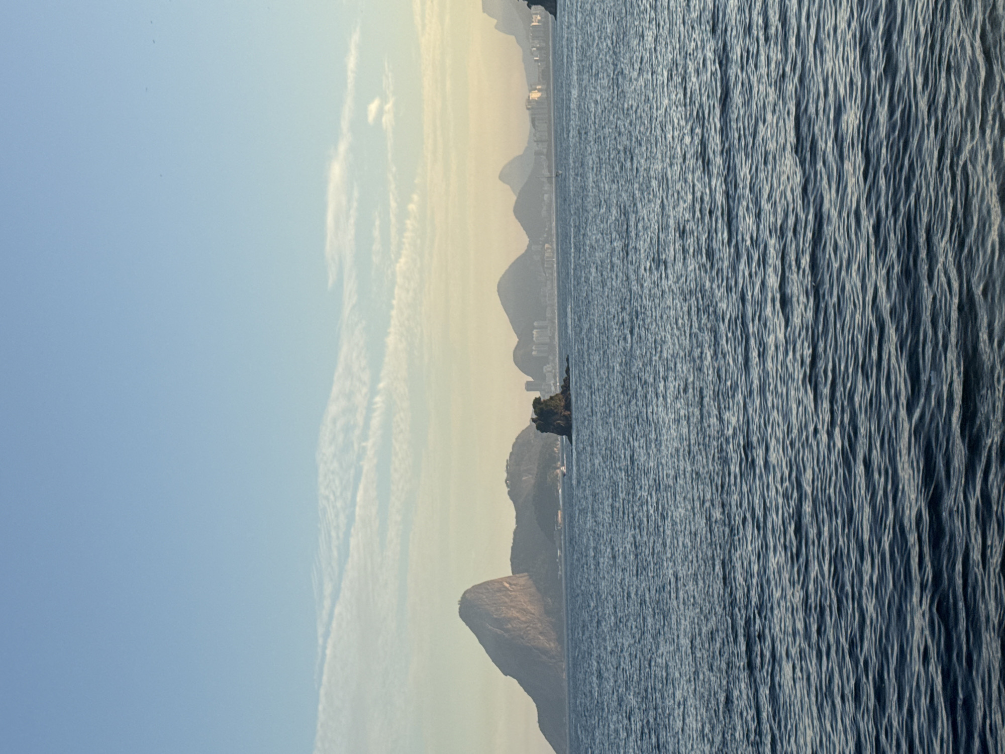The image captures a panoramic view of Guanabara Bay, looking towards Rio de Janeiro from Niterói, Brazil. The foreground is dominated by the rippling surface of the deep blue water. In the mid-ground, a distinctive rocky islet, likely Ilha das Cobras, stands prominently. Beyond the islet, the iconic Sugarloaf Mountain (Pão de Açúcar) rises dramatically, its steep, rounded peak a defining landmark. Behind Sugarloaf, a range of hills and mountains stretches across the horizon, dotted with the urban skyline of Rio de Janeiro, characterized by buildings of varying heights. The sky is a soft blue, with scattered wispy clouds tinged with the warm hues of either sunrise or sunset. The overall atmosphere is serene and picturesque, showcasing the natural beauty and urban landscape of the bay.