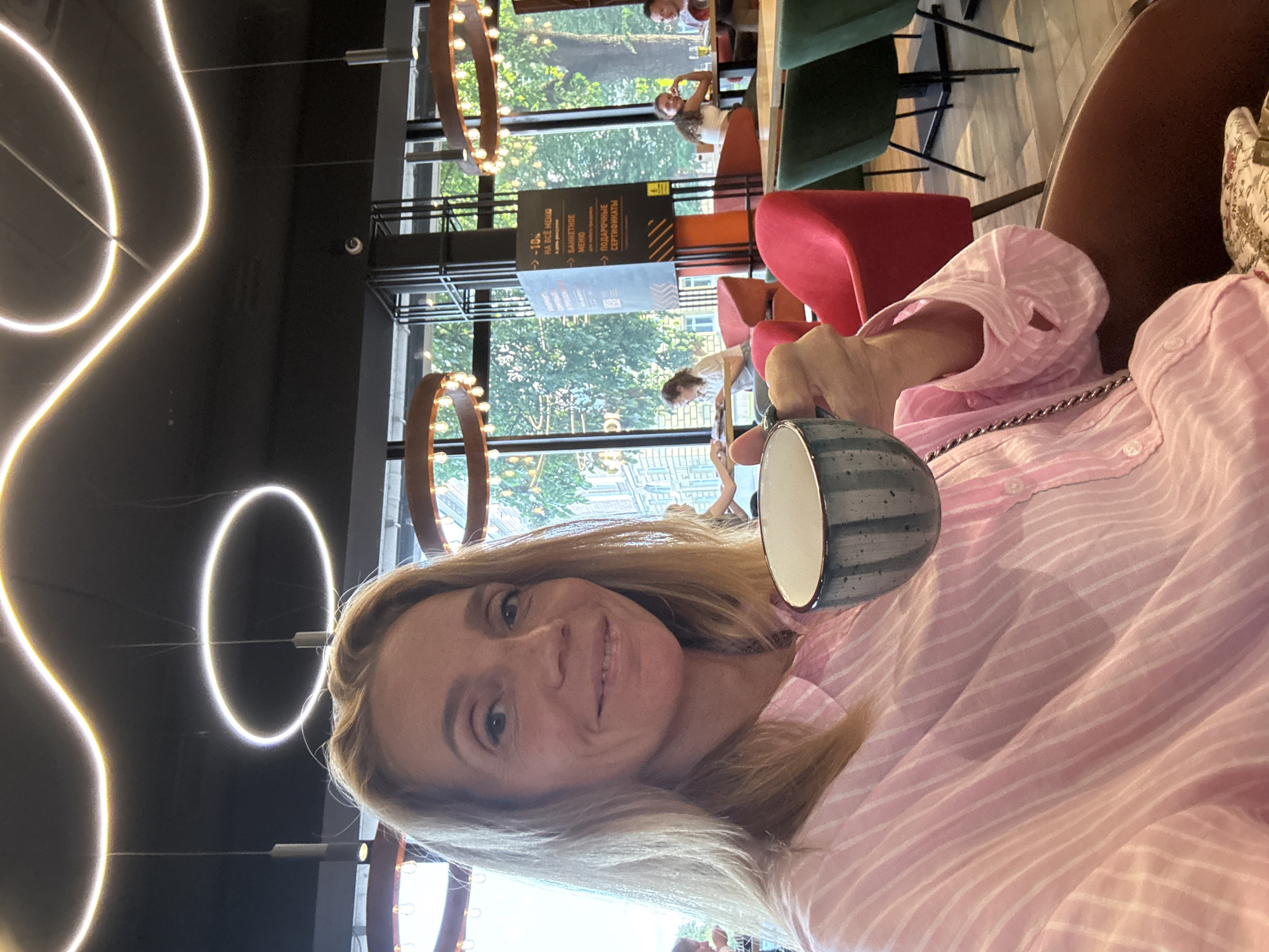 A blonde woman, appearing to be in her 40s, is taking a selfie while holding a cup with a blue and grey striped pattern. She is wearing a light pink and white striped button-down shirt and is seated in a brown armchair. The background suggests she is in a cafe or restaurant with large windows overlooking a tree-lined street, indicative of a public space in Veliky Novgorod, Russia. Modern light fixtures with wavy and circular designs are visible on the ceiling. Several other patrons are present in the background, some seated at tables. A sign with Russian text, including a discount offer of -10% and mentions of a banquet menu and gift certificates, is prominently displayed. The overall lighting is bright, suggesting it is daytime.