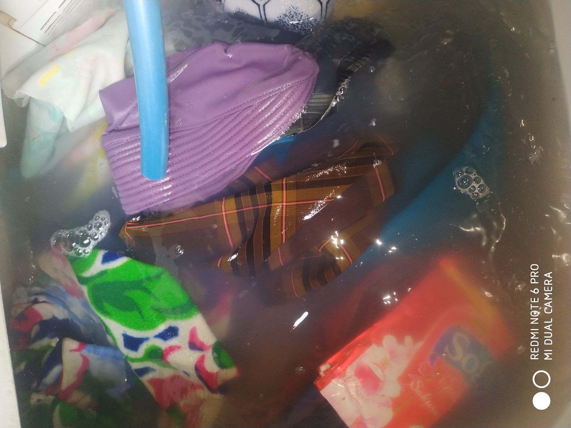 In Badas, Indonesia, a collection of laundry is submerged in murky water, likely in a washing basin. The items include a bright green and white patterned fabric, a purple pleated garment, a brown and pink plaid fabric, and a red and white packaged item with visible text. A blue pipe or hose is partially visible, submerged in the water and resting on the purple garment. Bubbles are present on the surface of the water, indicating agitation or the presence of soap. The overall environment appears to be a domestic setting, with the image capturing a close-up view of the laundry-submerged basin. The timestamp "REDMI NOTE 6 PRO MI DUAL CAMERA" is visible in the lower-left corner.