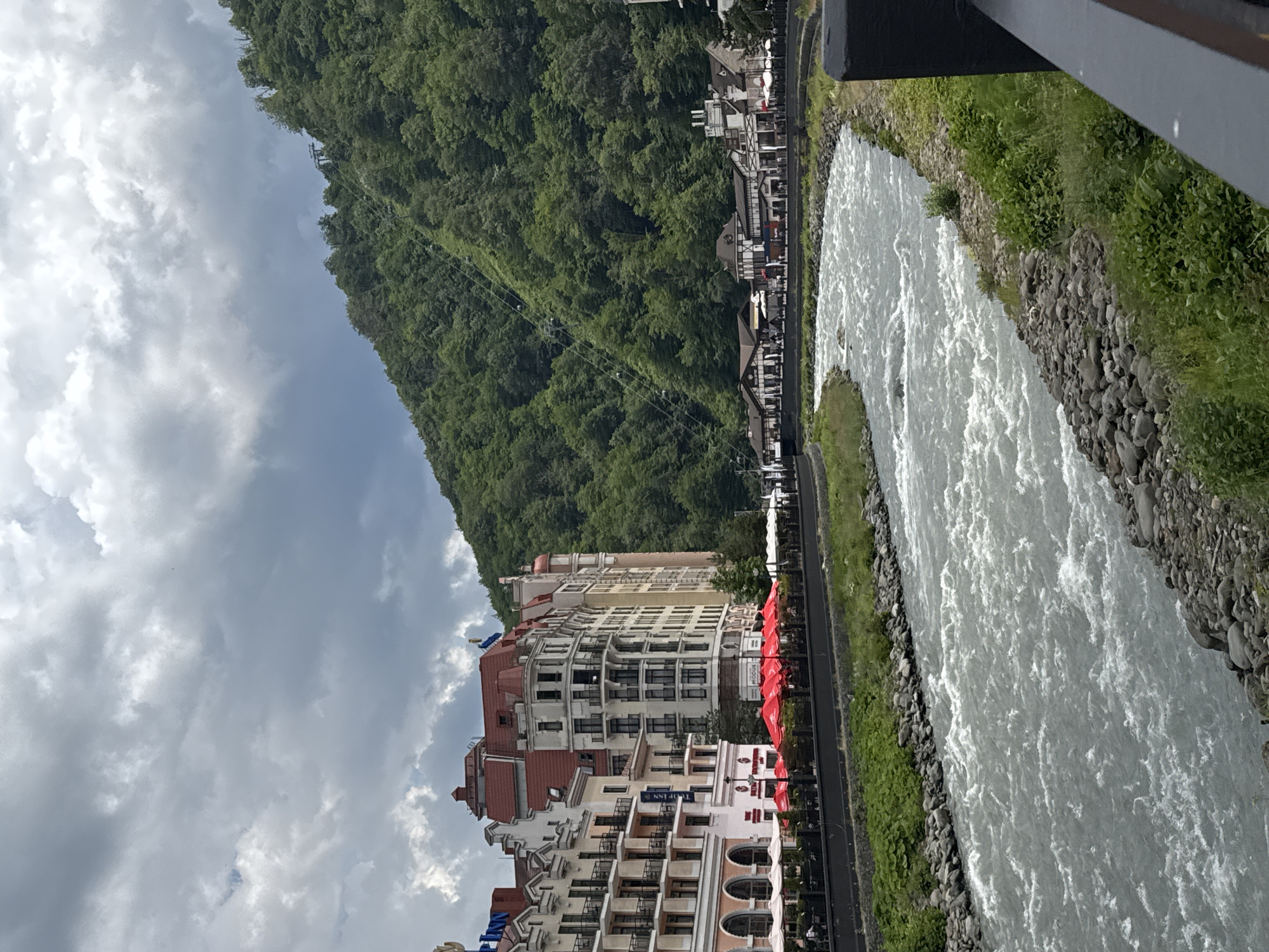 The image shows a river flowing through a town with European-style architecture. The buildings have red roofs and are situated along the riverbank. A lush, tree-covered mountain rises steeply behind the town, with a cable car system visible ascending its slope. The sky is overcast with dramatic clouds. The river is wide and appears to be flowing rapidly, with white water visible. The riverbanks are lined with rocks and green vegetation. There are outdoor seating areas with red umbrellas in front of some of the buildings. The scene appears to be a resort town, likely Estesadok, Russia, given the context.