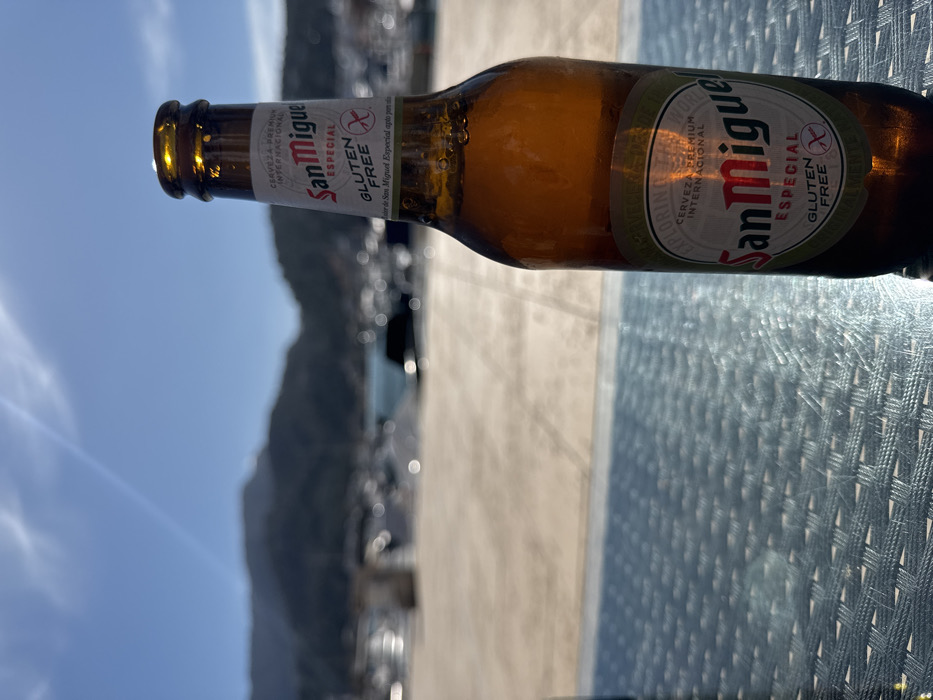 A brown glass bottle of San Miguel Especial Gluten Free beer, exhibiting condensation, stands prominently in the right foreground on a light blue woven table surface. The scene is located at a marina or port area within El Port, Spain. The midground depicts a light-colored paved waterfront, extending towards a body of water in the background. Numerous white boats and yachts are docked in the water, appearing out of focus. Beyond the marina, a range of dark, forested mountains is visible under a clear, bright blue sky with sparse, wispy clouds and a faint contrail. The lighting suggests full daytime. No individuals are present in the image.