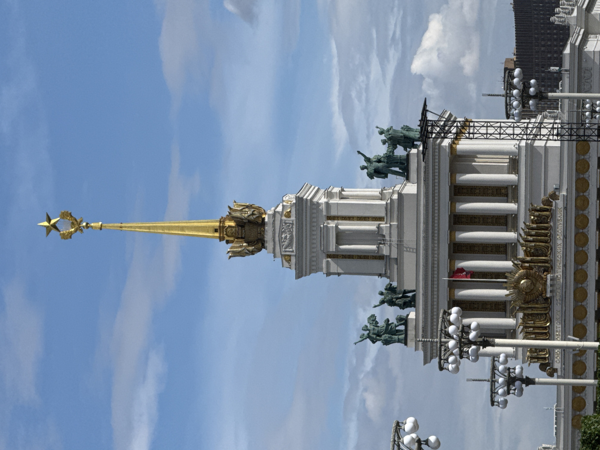 This media file, taken in Moscow, Russia, features a view of an ornate architectural building topped with a golden spire bearing a star. The building is adorned with classical columns, sculptural figures of people in dynamic poses, and decorative golden elements. Multiple lampposts with multiple spherical lights are visible in the foreground and midground, suggesting an outdoor setting. The sky is blue with scattered white clouds, indicating it is daytime. A portion of a modern, dark-colored building is visible in the background. Some text is visible on the facade of the main building, which appears to be in Cyrillic. The overall impression is one of grandeur and possibly a monument or a significant public building.