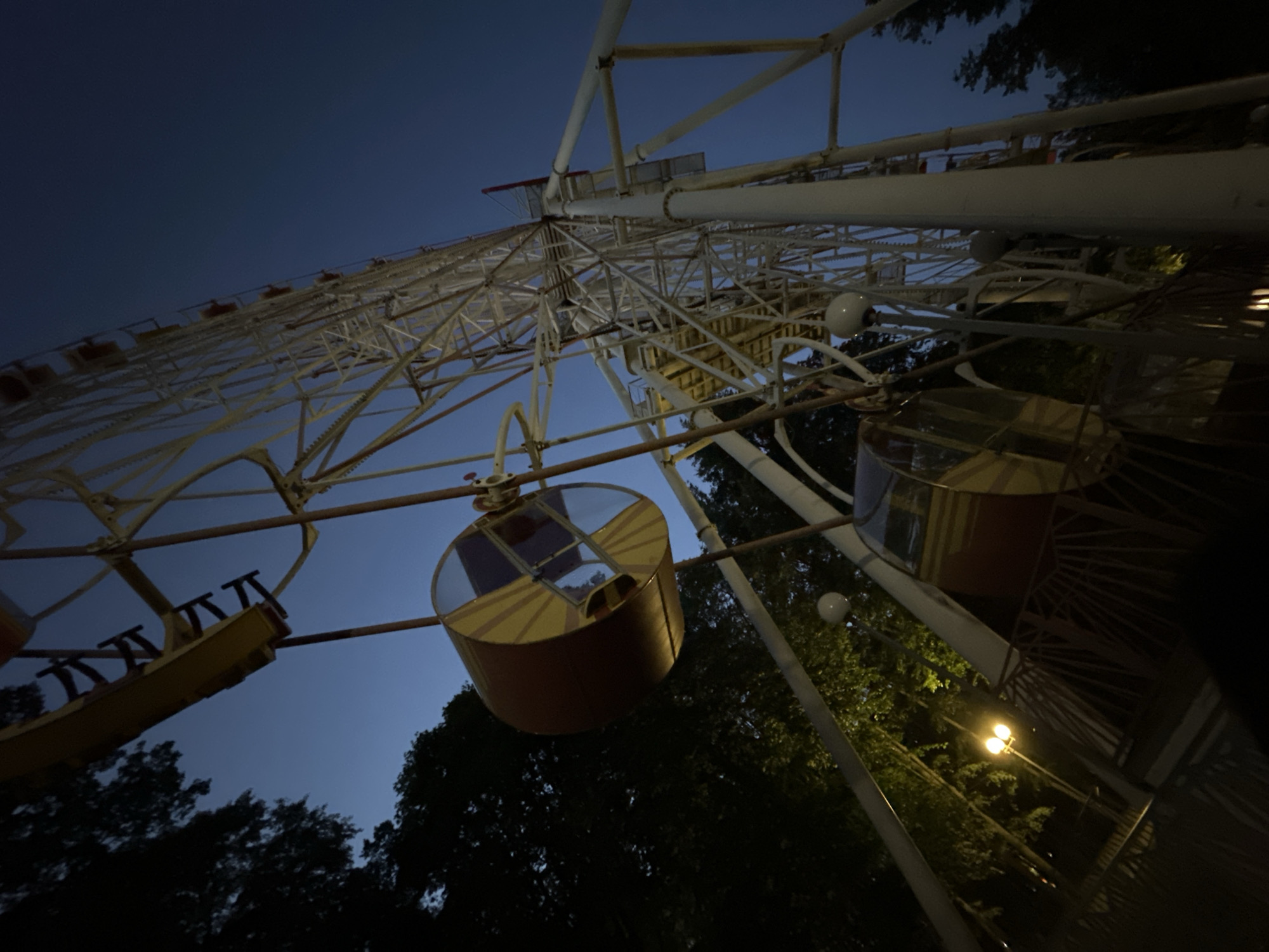 The image shows a Ferris wheel at dusk in an amusement park. The Ferris wheel is tall and white, with yellow and red cabins. Trees are visible in the background, and there are lights in the foreground. The sky is dark blue. The Ferris wheel is likely located in Gorky Park in Minsk, Belarus, as this park is known for its Ferris wheel.