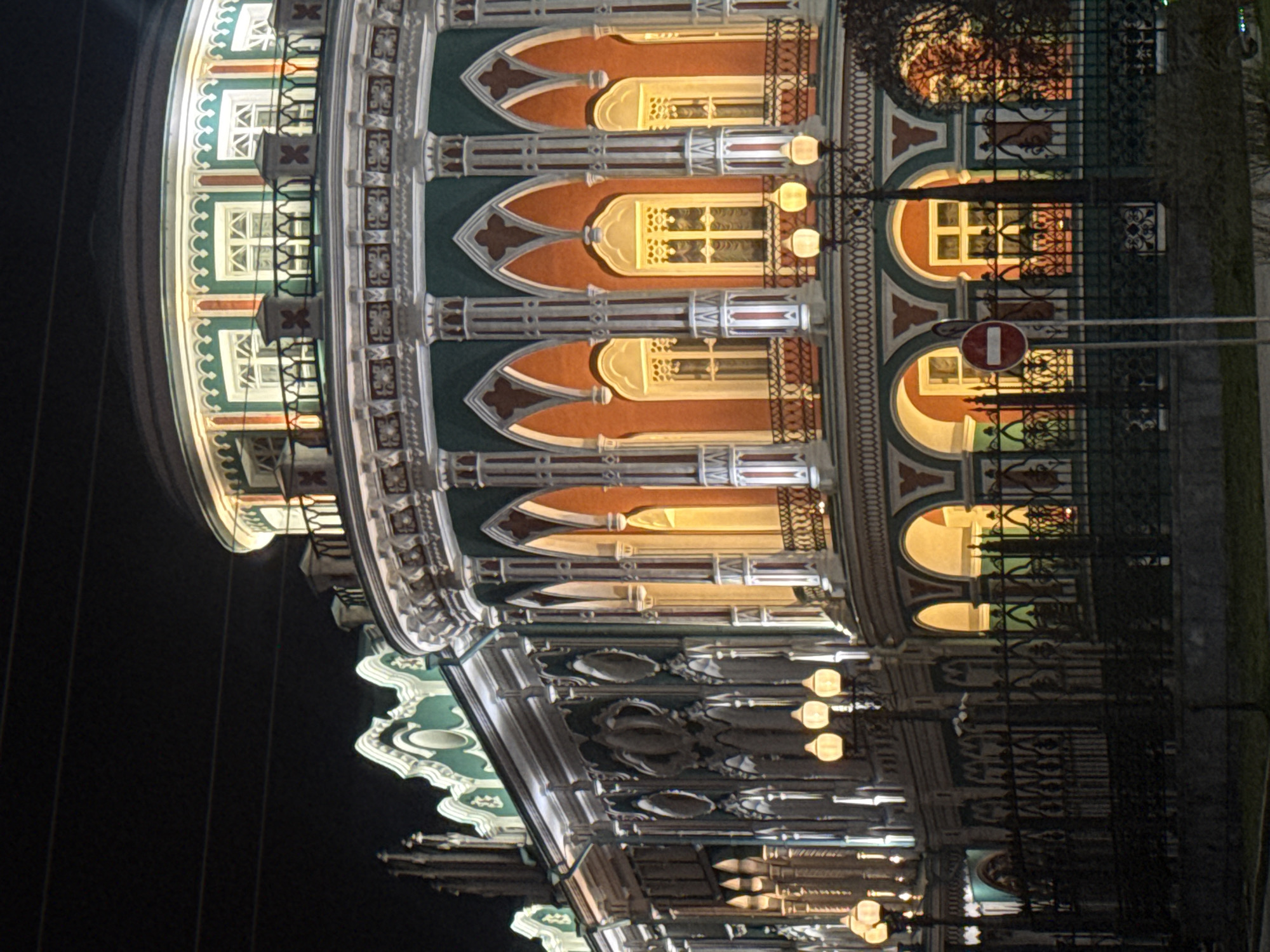 The image displays the illuminated facade of a grand, ornately decorated building at night, set against a dark sky. The architecture features an elaborate, possibly historic or revivalist style, characterized by numerous arched windows, decorative pillars, pointed arch motifs, and intricate patterns. The building's exterior is painted in a distinctive palette of deep green, warm terracotta/orange, and light grey/white, with detailing highlighted by bright lighting.
The scene is clearly nocturnal, with the building brightly lit. Warm yellow light emanates from within the many windows and from various exterior fixtures, casting a glow that emphasizes the architectural details. Balconies with ornate railings are visible in front of some windows.
In the foreground, a dark, intricate metal fence or gate runs along the perimeter of the building. A "Do Not Enter" traffic sign, depicted as a red circle with a white horizontal bar, is prominently visible on a pole near the fence, indicating an urban street environment.
No people or specific activities are depicted, focusing solely on the majestic edifice. The building's detailed and repetitive decorative elements, including its cornices and stylized patterns, are a notable feature. The overall impression is one of a richly detailed and well-preserved structure, possibly in Yekaterinburg, Russia, as suggested by the context. No explicit text is visible, beyond the universal symbol on the traffic sign.