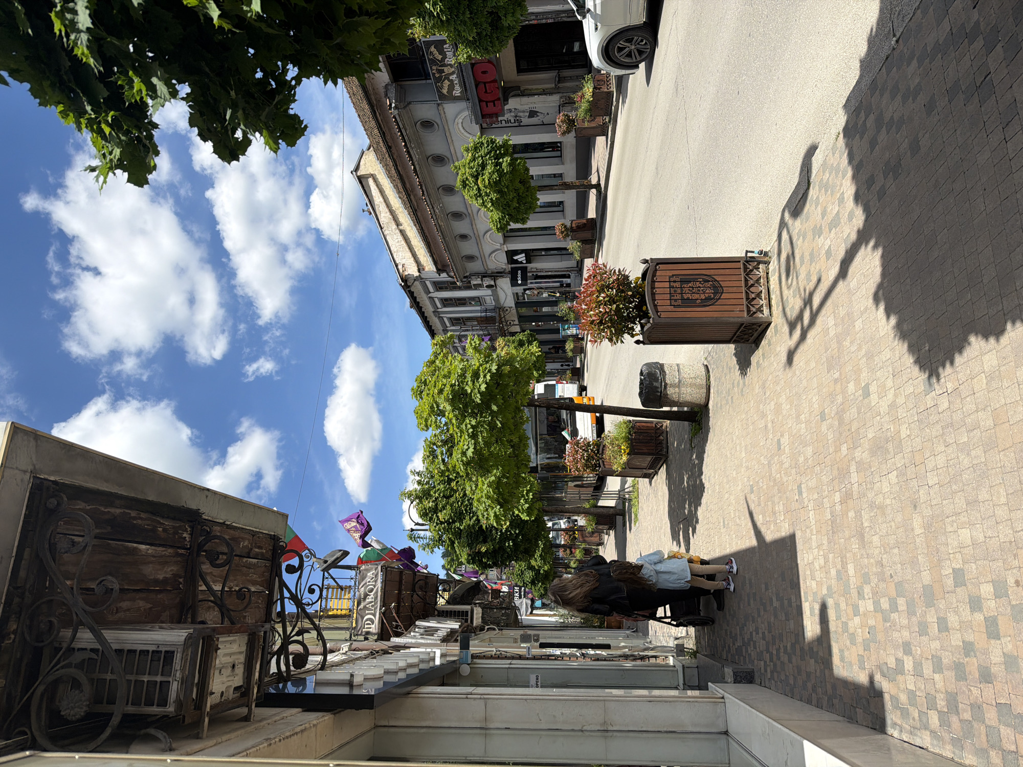 The image depicts a street scene in Veliko Tarnovo, Bulgaria. The camera angle is looking down the street and slightly to the left.
People are present, two girls with long hair, one in a black jacket, and another next to her in light-colored clothing, are seen walking down the pedestrian walkway.
The environment consists of a narrow street paved with stones, buildings lining both sides, adorned with shops, restaurants, and decorative elements. Planters with small trees and flowers are placed along the street. The sky is visible, with blue patches and scattered clouds. There are signs in Bulgarian visible on the buildings. The buildings show various styles of architecture. A car is parked on the side of the street.