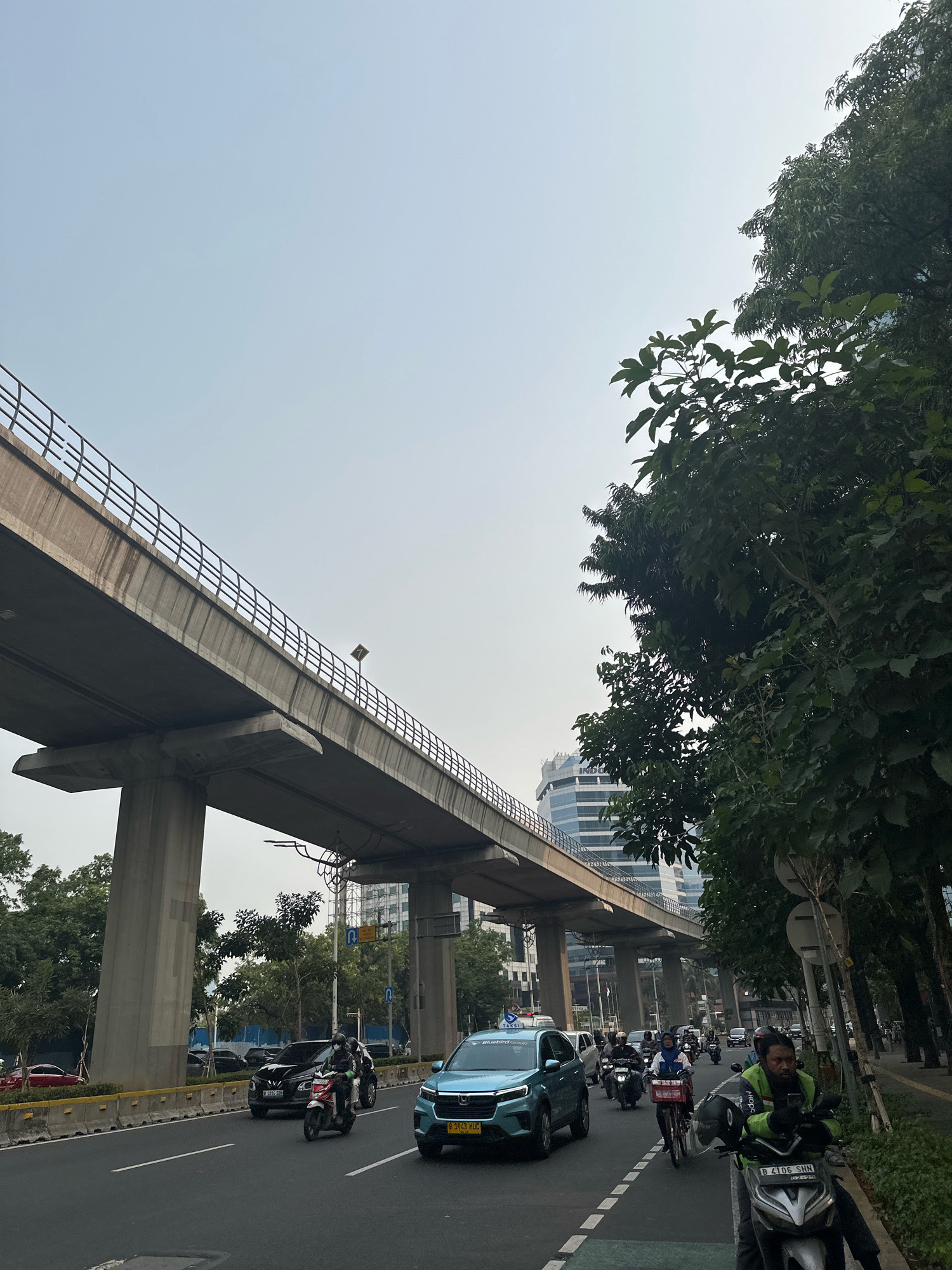 In the city of South Jakarta, Indonesia, a photograph captures a street scene beneath an elevated railway line. The sky is a pale, hazy blue. The concrete structure of the railway, supported by large pillars, dominates the upper left of the frame. Trees with dense green foliage line the right side of the road, partially obscuring modern buildings in the background.
The road itself is a multi-lane thoroughfare with several vehicles. A light blue SUV is in the foreground, followed by a line of motorcycles. One motorcycle rider, wearing a bright green jacket and a helmet, is prominently featured on the right. Other vehicles, including cars and motorcycles, are seen moving in the same direction. Traffic appears to be moderate. A pedestrian walkway is visible along the right side of the road, bordered by greenery. The overall impression is one of urban transit and daily life in a bustling Indonesian city.