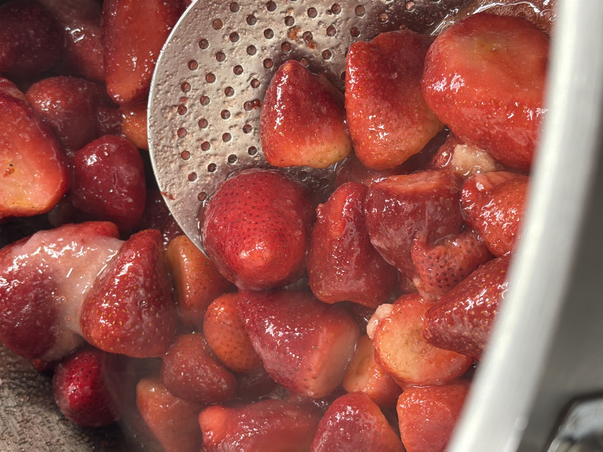 This close-up image captures a vibrant collection of whole and halved strawberries being cooked in a pot or deep pan, likely for a jam, compote, or sauce. The strawberries are a rich, glossy red, appearing softened and glistening from the surrounding sweet liquid, which is a reddish syrup or their own released juices. Some berries show lighter, almost white or pale pink sections, indicating areas of less ripeness or cut surfaces.
A metallic slotted spoon, with numerous small, circular perforations, is partially submerged among the fruit, suggesting it has been used to stir or lift the strawberries. The texture of the spoon's surface is visible, showing small indentations or wear. No people are visible in the frame, focusing entirely on the culinary process. The lighting is bright and even, highlighting the vibrant colors and the sheen on the fruit. There is no visible text in the image. The scene conveys a sense of home cooking or food preparation. This activity takes place in Belgrade, Serbia.
