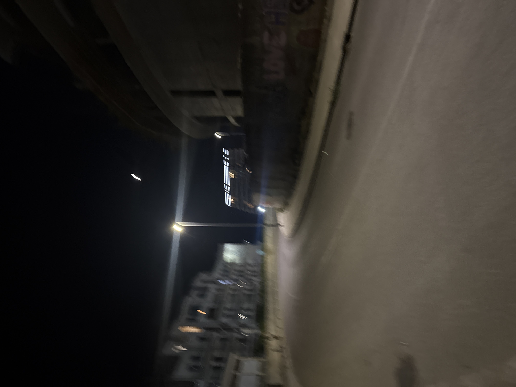 The image depicts a dimly lit street scene at night in Boreti, Montenegro. A curved road leads away from the viewer, with a sidewalk and a low wall bordering the right side. Buildings are visible in the background on the left, appearing as blurred structures. A street lamp illuminates the area with a bright light. Graffiti, partially legible as "LOVE HE," is spray-painted on the wall on the right. The overall impression is one of a quiet, urban environment after dark.