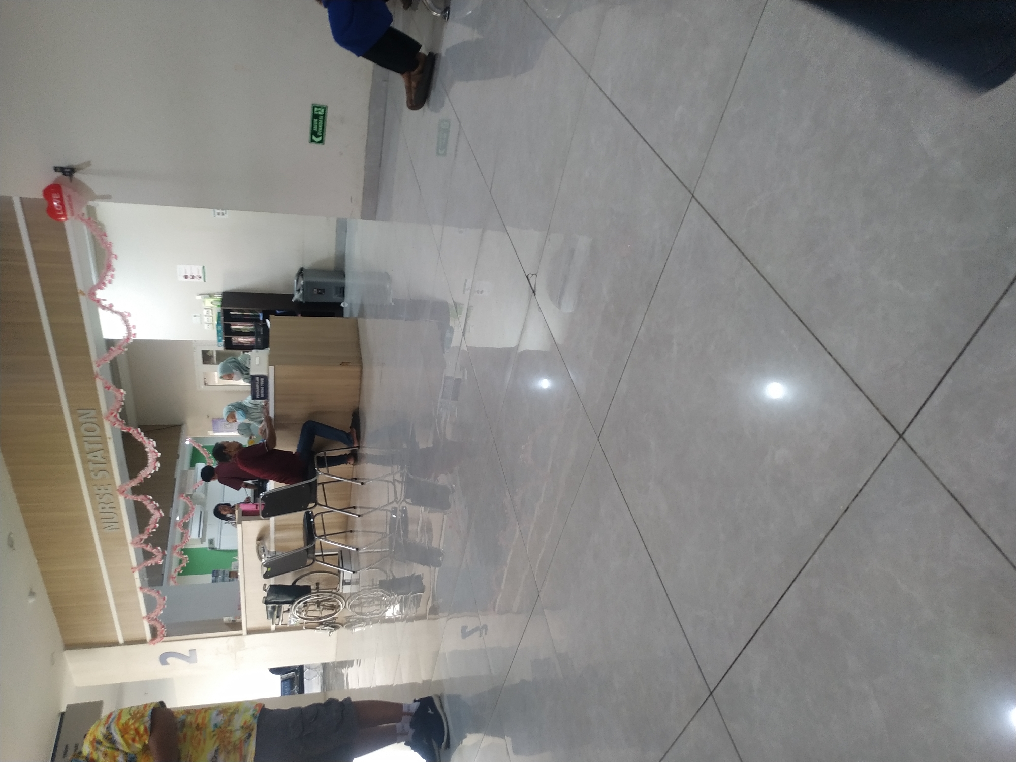 The image depicts the interior of a healthcare facility, likely a clinic or hospital reception area, in Mojosongo, Indonesia. The "NURSE STATION" sign is prominently displayed above a counter where two individuals are interacting. One person, wearing a maroon shirt and jeans, is seated at the counter, appearing to be a patient or visitor. Another individual, wearing a light blue headscarf and possibly a medical uniform, is behind the counter, possibly a nurse or receptionist. To the left of the counter, a wheelchair is parked. The floor is tiled with a light grey material, reflecting the overhead lighting. A red heart-shaped balloon with the word "LOVE" is attached to the wall on the right. Further to the right, a green exit sign with "JALUR EVAKUASI" (evacuation route) is visible. A person in a blue garment is partially visible on the far right, sitting on a chair. The overall environment appears clean and organized, with a light-colored wall and wood-paneled accents.