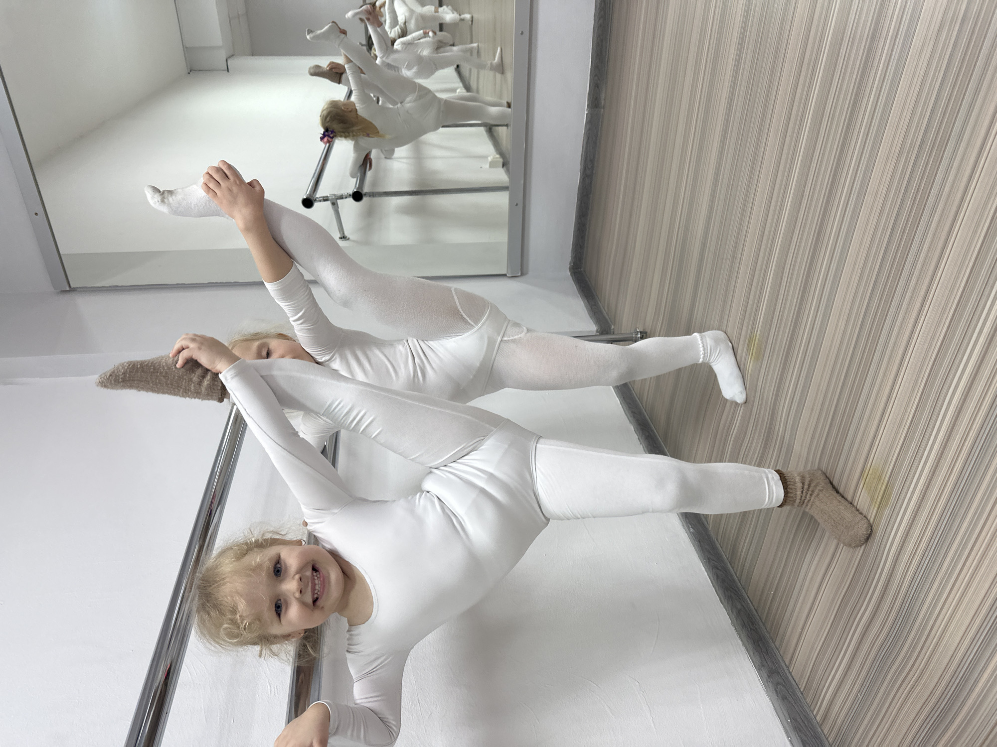 Two young girls in white leotards are performing splits in a dance studio. The girl in the foreground has blonde, curly hair, a tan sock on one foot, and is smiling. She is holding her leg up towards her head. The girl in the background also has blonde hair, white socks on her feet, and is holding her leg up towards her head. They are holding onto a ballet barre. A mirror on the wall reflects the girls and the barre. The studio has a wood-look floor and white walls. The location is likely in Votkinsk, Russia.