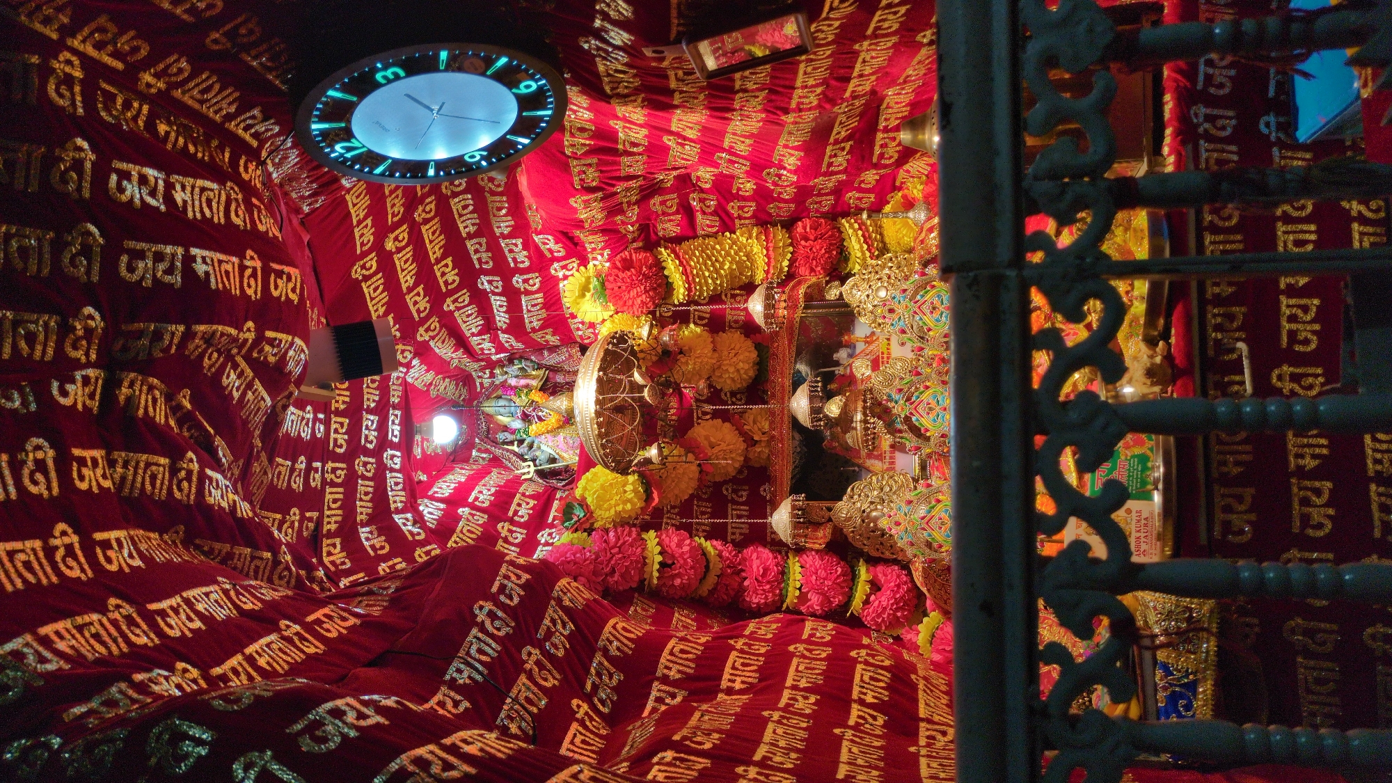 This vibrant indoor image captures a richly decorated shrine or temple, likely dedicated to a Hindu goddess, in Bhagsu, India. The space is adorned with draped red fabric featuring repeated golden Devanagari script that reads "जय माता दी" (Jai Mata Di), a devotional chant meaning "Victory to the Mother Goddess." Numerous garlands of bright red and yellow flowers are strung across the ceiling and around ornate golden decorative elements, which likely form part of an altar or deity's adornment.
On the upper left, a circular wall clock with a glowing blue rim displays the time as approximately 9:45. Artificial lighting, including bright bulbs, illuminates the sacred space, suggesting a nighttime or low-light setting. To the right, a dark, intricately designed metal railing partially obstructs the view. A small reflective surface or framed picture is also visible amidst the decorations.
Visible text on a small sign near the center includes "ASHOK KUMAR & JAURA" and "KIL DHARAMSHALA", possibly indicating donors or a nearby establishment. The overall atmosphere is one of reverence, festivity, and spiritual devotion, characterized by the rich colors and religious symbolism common in Hindu places of worship.
