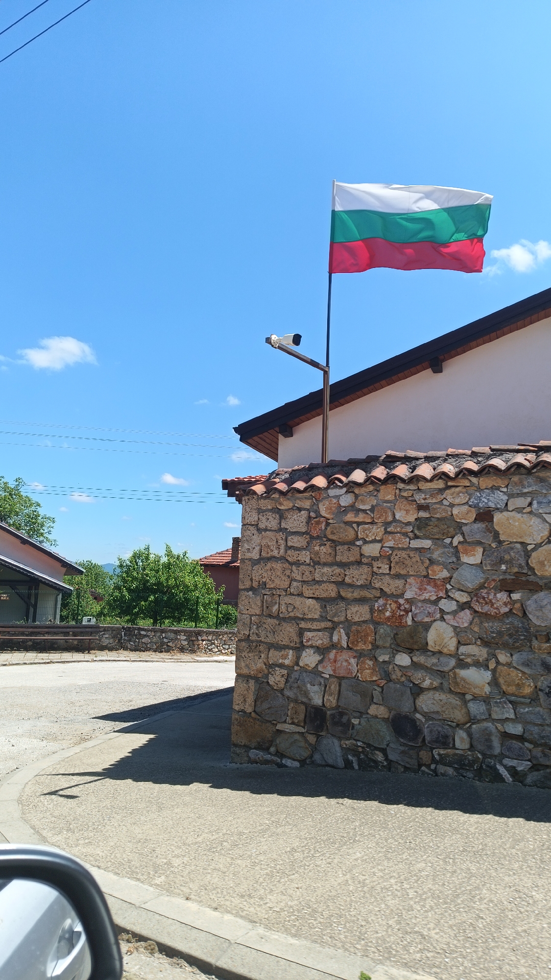 The image depicts a street scene in Gornoslav, Bulgaria. A stone wall with terracotta tile coping stands on the right side of the frame. A Bulgarian flag waves atop a metal pole attached to a white building behind the wall. A security camera is mounted on the pole below the flag. The sky is blue with scattered clouds. The street is paved and curves to the left. Other buildings and trees are visible in the background. The side mirror of a vehicle is partially visible in the lower-left corner.