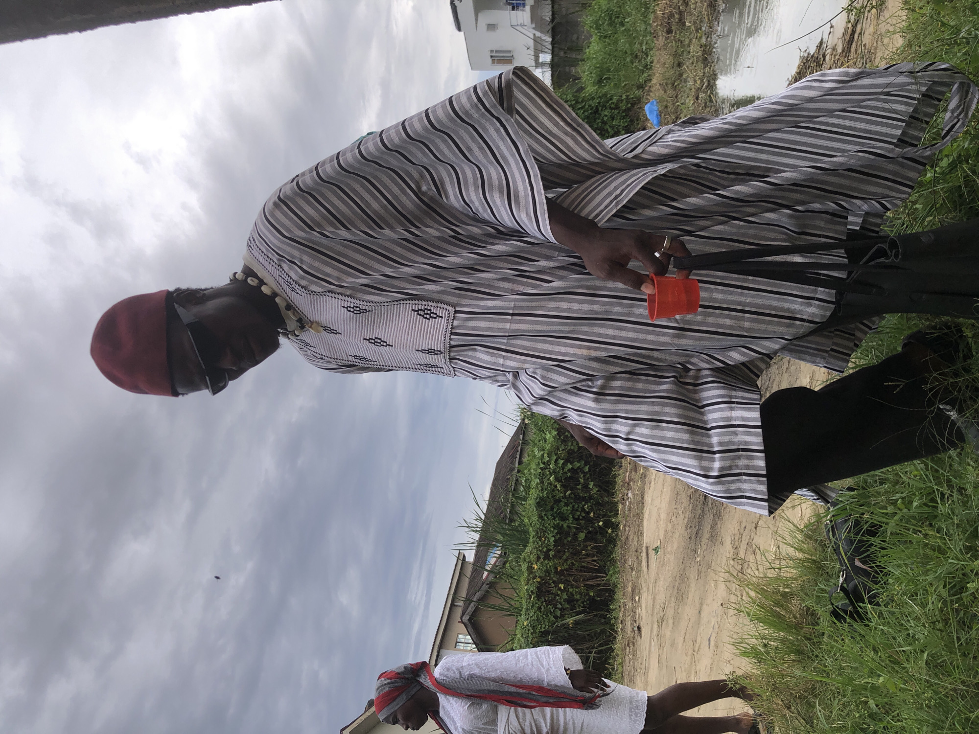 In Ado, Nigeria, under an overcast sky, a man in traditional attire, including a red cap, dark sunglasses, and a striped robe with a beaded necklace, stands holding a small red cup. He wears a ring on his left hand. To his left, a woman in a white dress and headscarf with red and grey accents walks away from the camera. The ground is a mix of dirt and grass, with a muddy puddle and some construction in the background. A black bag is partially visible in the grass near the woman's feet. The scene appears to be an outdoor gathering or event.