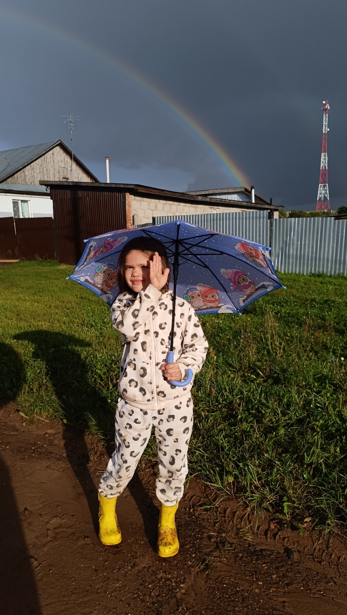 A young girl stands in a rural outdoor setting with a rainbow in the background. She is holding an open blue umbrella with owl designs on it. She is wearing a leopard-print tracksuit and yellow rain boots. The ground is muddy and grassy. In the background, there are buildings, a metal fence, and a telecommunications tower. The sky is dark with a rainbow arc. The scene appears to be in Karsha, Russia.