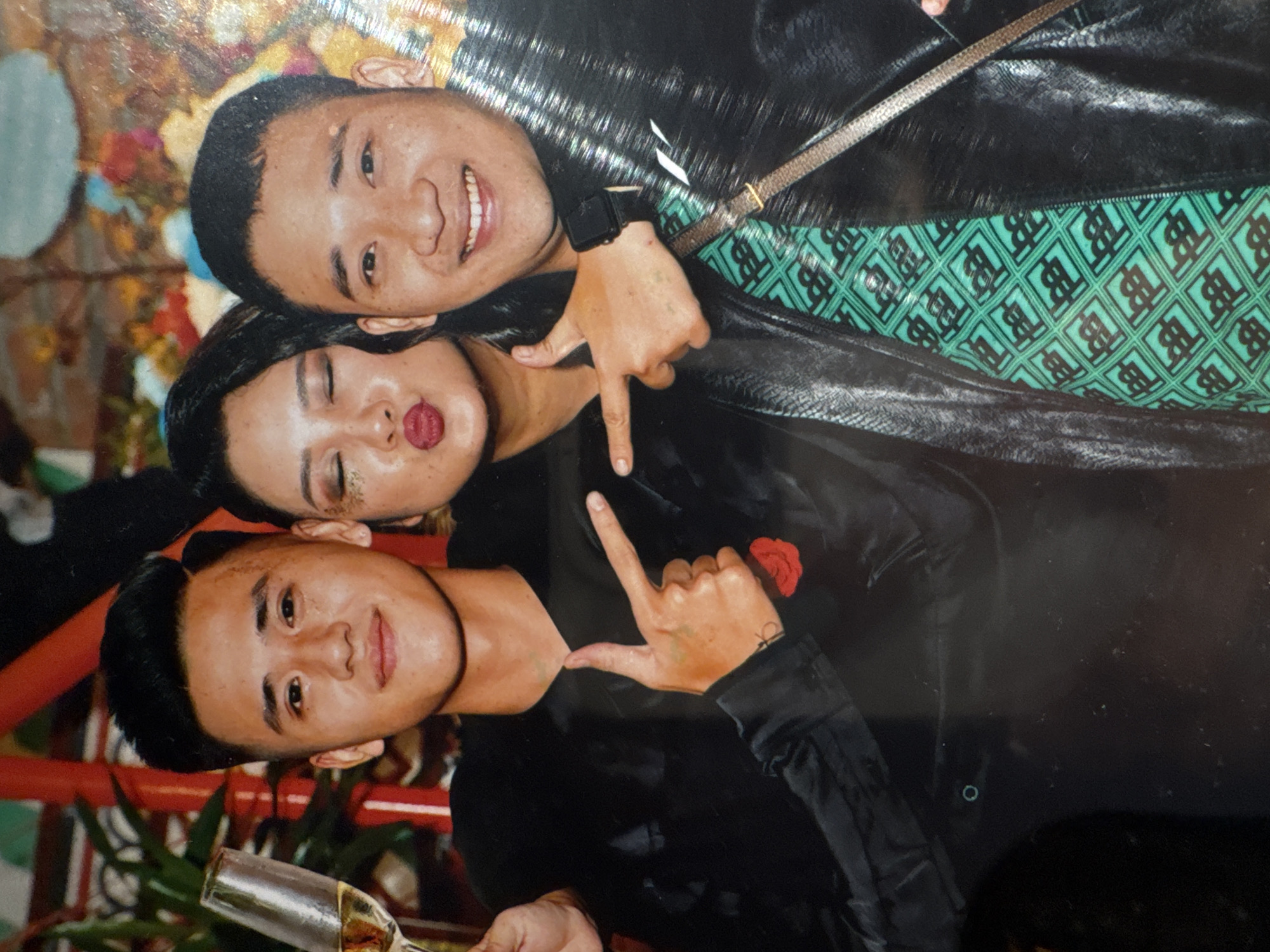 The image shows three young adults posing for a photo. The person on the left, a young man with short black hair, is holding a champagne flute and smiling. The person in the middle, a young woman with dark hair, is making a pouty face with glitter on her eyelids. The person on the right, a young man with short black hair, is smiling broadly. All three are wearing dark clothing. The background appears to be an indoor setting with some colorful decorations. The image quality is somewhat grainy and appears to be a scanned photograph. There are no discernible landmarks or details that would place this image specifically in Hanoi, Vietnam, or any other city.