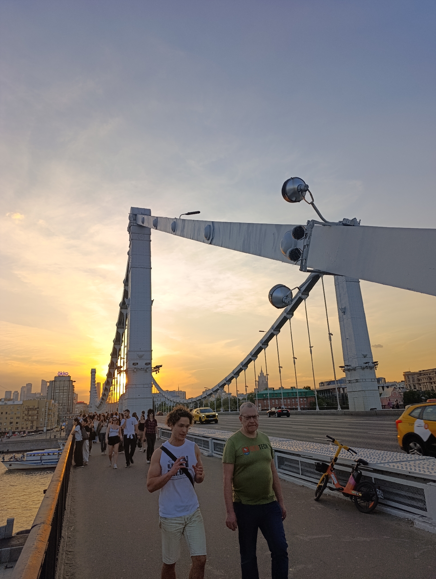 This image depicts the Krymsky Bridge (Crimean Bridge) in Moscow, Russia, during sunset or golden hour. The view is looking generally southeast, with the setting sun low on the horizon, casting warm, orange light across the sky and cityscape.
On the left, the Moscow River is visible with a white tour boat moving away from the viewer. Along the riverbank, various buildings of Moscow's city center are present, including a structure with a prominent "ökko" sign. Distant skyscrapers, likely part of Moscow City, are silhouetted against the sky. A tall, spired building, characteristic of Stalinist architecture, is also visible further in the background.
The foreground shows the pedestrian walkway of the bridge. Two men are prominent:
1. On the right, an older man with glasses and a green t-shirt stands facing the camera, wearing blue jeans.
2. In the center, a younger man with curly brown hair, wearing a white tank top and light shorts, walks towards the left, appearing to hold a small device in his hands.
Behind these two individuals, a crowd of people walks along the pedestrian path, generally heading towards the sunlit western end of the bridge.
To the right of the pedestrian walkway is the vehicle roadway of the bridge, with several cars, including yellow taxis, driving in both directions. Parked against the railing on the pedestrian side is an orange and yellow shared electric scooter or bicycle.
The distinctive white towers and suspension cables of the Krymsky Bridge are prominent features, with several large, spherical light fixtures mounted on the bridge's structural elements. The sky exhibits a gradient from deep blue at the top to vibrant orange and yellow near the horizon, with scattered clouds.