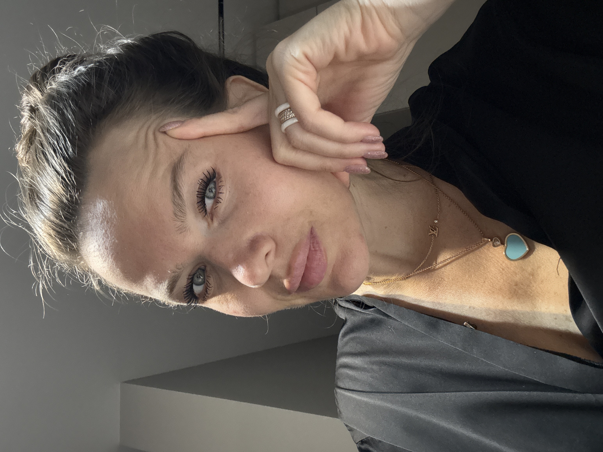 A woman with fair skin, light blue eyes, and dark hair pulled back in a loose bun, poses for a selfie. She is wearing a black top that appears to be silk, and a gold chain necklace with a heart-shaped pendant made of light blue stone. She has a large ring on her left ring finger, comprised of white bands and a sparkling band. Her hand is raised to her temple. The background shows a plain white wall. There is no indication of this being in Khimki, Russia.