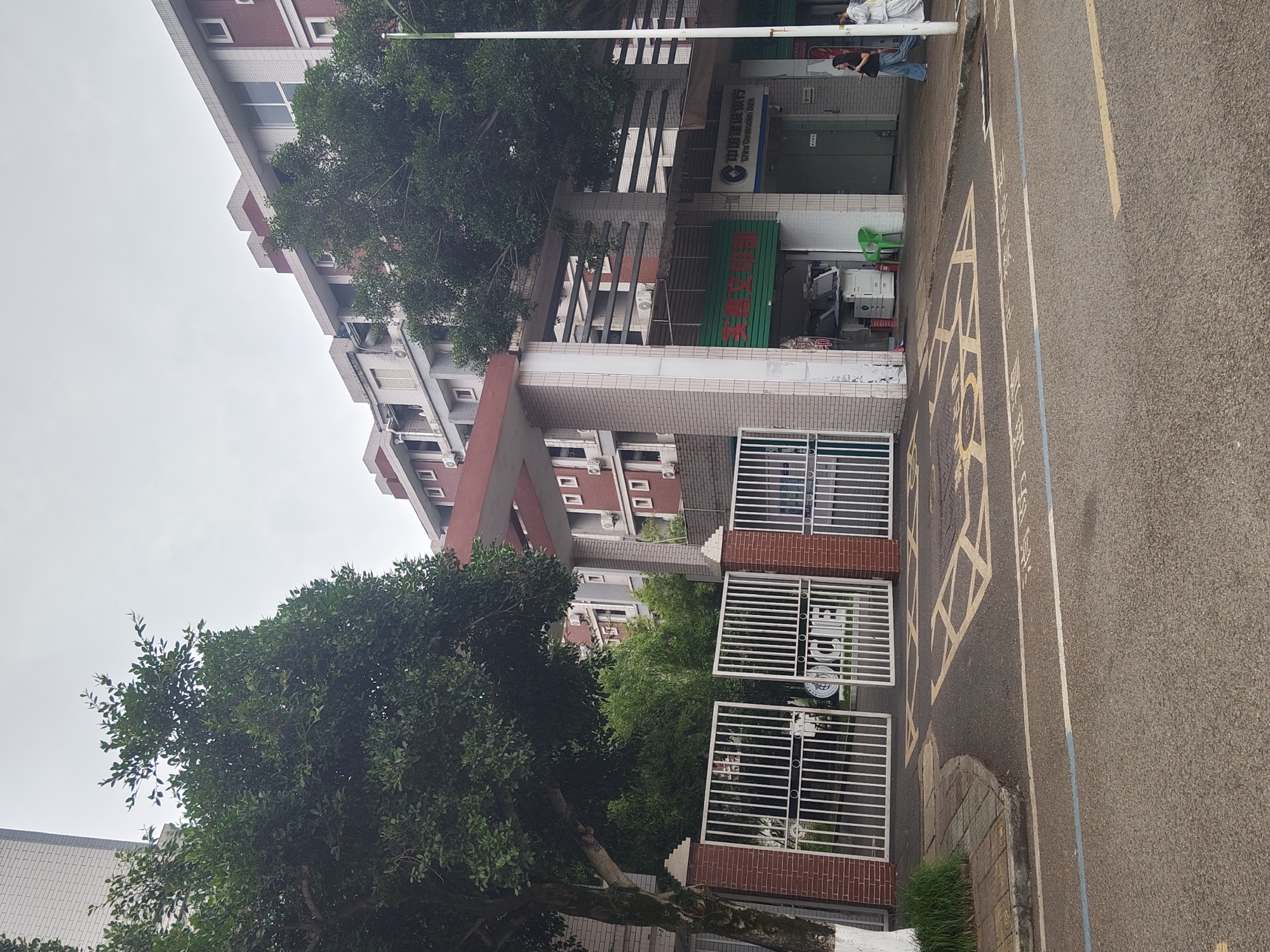 The image shows a street scene in the Chongqing High-tech Zone, China, with a building complex on the right and a street in the foreground. The building is a multi-story structure with a red brick facade on the upper floors and light-colored bricks on the lower levels. There are numerous windows and air conditioning units visible on the building. In front of the building, there are two white metal gates, partially open, leading into a courtyard or walkway. A tree with dense green foliage stands to the left of the gates, and another large tree is positioned to the right, partially obscuring the building. On the ground floor of the building, there are storefronts, one of which appears to be a printing shop with photocopiers visible inside. Another storefront has signage for "China Construction Bank." The street in the foreground is paved, with yellow traffic markings, including a crosswalk. Two people are walking on the sidewalk in front of the bank. The sky is overcast.