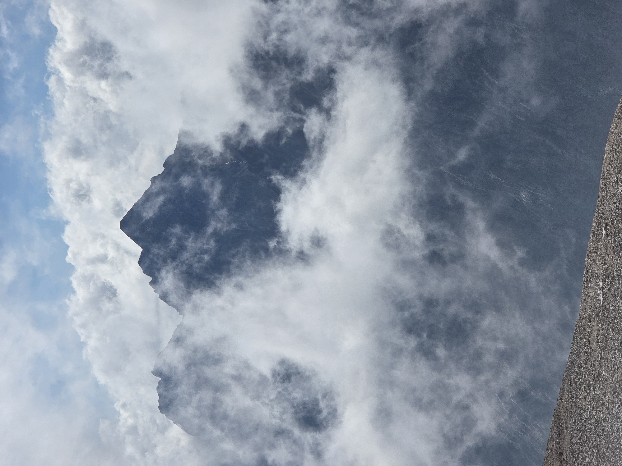 The image is a photograph of a mountain peak shrouded in clouds, with a foreground of rocky terrain. The sky above is partly cloudy. The image is taken from a high vantage point, looking upwards towards the mountain. There are no people or man-made objects visible in the scene. The environment is mountainous and natural, with the clouds creating a dramatic and atmospheric effect. The location is not Garmisch-Partenkirchen, Germany.