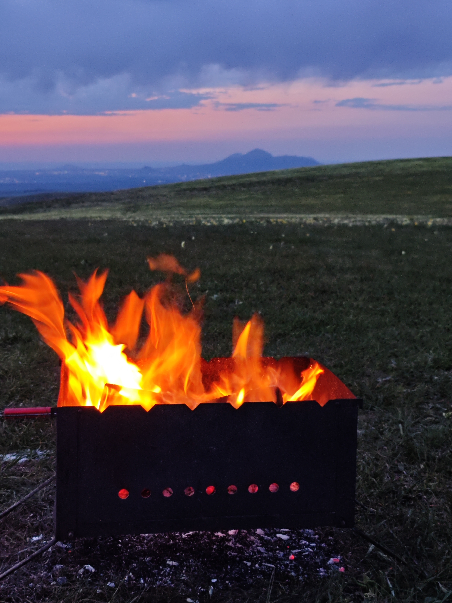 A grill with a roaring fire is centered in a grassy field in Unknown, Unknown. The sky is a mixture of deep blue and dusky pink. There is a gentle, green hill in the midground. A smattering of lights in the far distance could be a city. A mountain range is barely visible on the horizon.