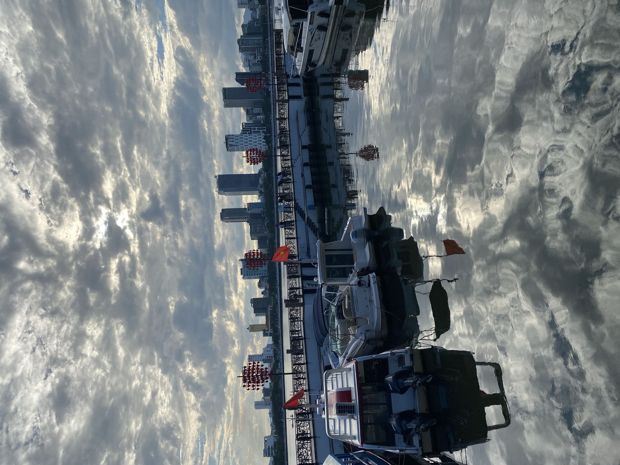 A cloudy sky with dramatic, illuminated clouds hangs over a city skyline featuring modern skyscrapers. Below the skyline, a pier with decorative red lanterns and black railings is visible, with people strolling along it. The pier extends over a calm body of water where several boats are docked. In the foreground, three boats are prominently featured: two motorboats with twin outboard engines and one larger white motorboat. The reflections of the sky, city, pier, and boats are clearly visible on the water's surface. The scene is likely captured at dusk or dawn due to the lighting and cloudy conditions. The presence of Vietnamese flags on some of the boats suggests the location is in Vietnam, specifically Đà Nẵng, Vietnam, given the characteristic skyline.