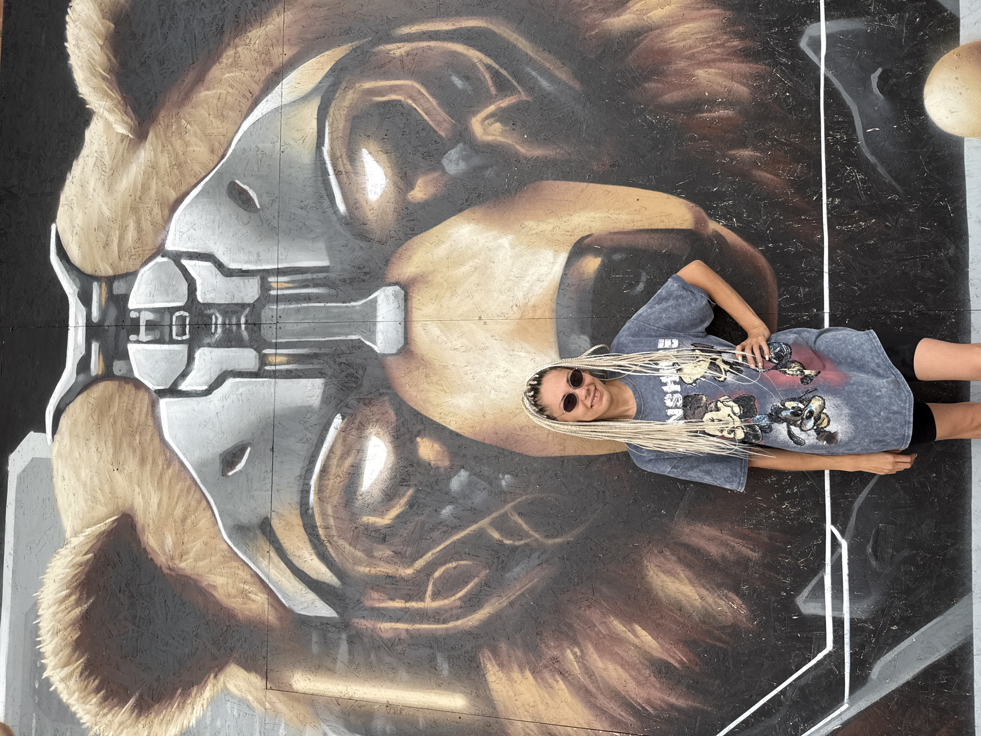 In the city of Rusanovo, Russia, a woman with blonde braided hair and dark sunglasses poses in front of a large mural of a bear's head. The bear's head is depicted with a metallic helmet covering its face, creating a cybernetic appearance. The mural is painted on a rough, wooden surface, possibly a wall or fence. The woman is wearing a dark grey t-shirt with a graphic print and black shorts. Her left hand rests on her hip, and she is smiling at the camera. The background mural features the bear's face with glowing yellow eyes, and the texture of the wood is visible beneath the paint. The overall scene is vibrant and artistic, with the woman and the mural being the primary subjects.