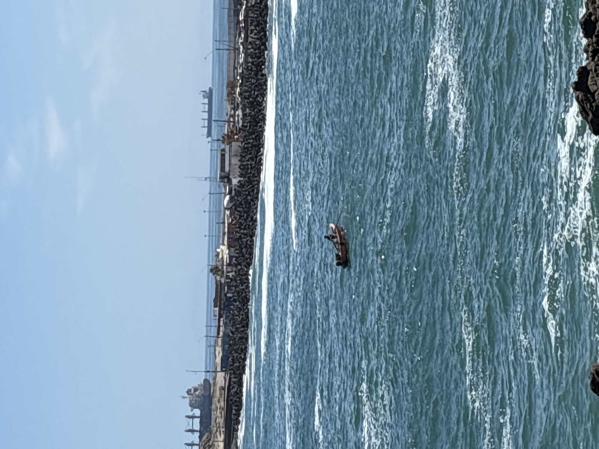 This image captures a bright daytime scene along the coast of Arica, Chile, showcasing a small boat on the ocean near a breakwater.
The central subject is a lone individual in a dark, small boat, possibly a fishing dinghy, situated in the blue-green, choppy ocean water. The person appears to be facing away from the viewer, seemingly engaged in an activity like fishing or navigating the boat.
To the left of the boat, a long, dark breakwater or pier, constructed from irregular blocks (likely tetrapods or large rocks), extends into the distance. Several slender poles, which could be fishing rods or small utility poles, are visible along this structure. In the far background, beyond the breakwater, a faint horizon meets a clear, light blue sky, with some indistinct structures or larger vessels visible on the distant water.
On the far right edge of the frame, a dark, rugged rocky formation, possibly a cliff or natural coastline, is partially visible. The ocean surface is textured with numerous waves and whitecaps, suggesting active water movement. The sky is largely clear and bright, indicating good weather conditions. No text is visible in the image. The overall scene depicts a calm coastal environment with typical maritime activity.