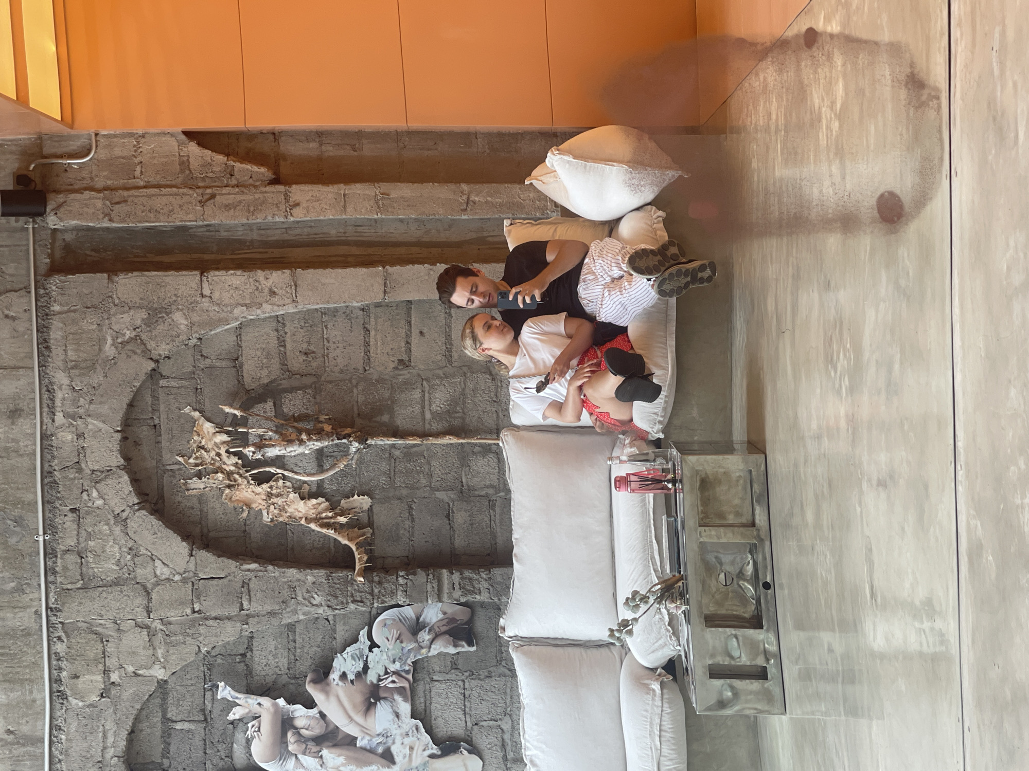 Two individuals, a man and a woman, are seated on a white sectional couch in what appears to be a cafe or lounge area. The wall behind them is constructed of rough, grey stone blocks, with two archways visible. To the left, a collage-like artwork featuring nude figures is affixed to the wall. On the right side of the image, a portion of an orange wall is visible. The floor is a polished, light-colored concrete.
The woman, on the left, is wearing a white t-shirt and a red and white checkered skirt. She is holding a smartphone in her right hand and her left arm rests on her lap. Her head is tilted down, and she is looking at her phone.
The man, on the right, is wearing a black t-shirt and striped pajama pants. He is also holding a smartphone and appears to be looking at it. His right arm is around the woman, and his left leg is extended, with his feet resting on the floor.
In front of the couch, there is a rectangular, metallic coffee table with various items on it, including a small bouquet of flowers, a pink diffuser bottle, and a book. The overall ambiance suggests a relaxed and contemporary setting. The scene is likely located in the city of North Kuta, Indonesia, given the context provided.