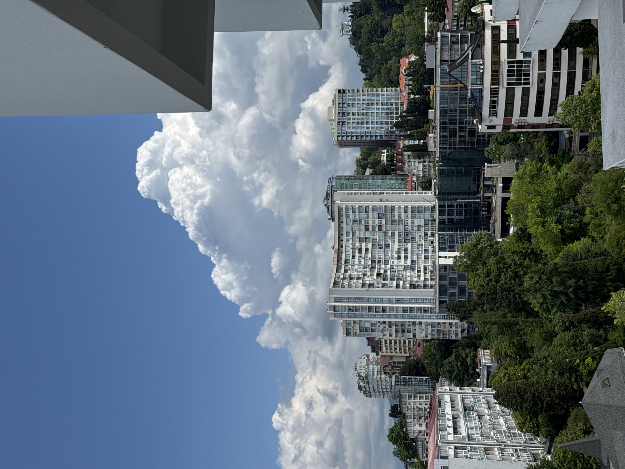 This image captures a view of the city of Sochi, Russia, with a focus on its modern architecture nestled amidst lush greenery and a dramatic sky. The foreground features a portion of a white building's structure, suggesting the vantage point is from a balcony or rooftop.
Dominating the mid-ground are several tall, multi-story buildings, indicative of urban development. One prominent white building, with a curved facade, is adorned with numerous windows and balconies, reflecting the bright sunlight. Its rooftop sports signage. Adjacent to it are other high-rise structures of varying designs, some with sleek, glass exteriors, and others with more traditional apartment block appearances. Construction is visible on some buildings, with scaffolding and netting indicating ongoing development.
The buildings are situated on a hillside, with dense, dark green trees interspersed throughout the urban landscape. The trees are a mix of coniferous and deciduous varieties, creating a rich, verdant tapestry that contrasts with the concrete and glass of the city.
The background is dominated by a vast expanse of blue sky, dramatically punctuated by large, voluminous white cumulus clouds. These clouds are so substantial they appear to be a significant feature of the landscape, casting subtle shadows on the scene below. In the far distance, on a higher slope, a white structure, possibly a cable car station or observation tower, is visible. The overall impression is one of a contemporary city that has successfully integrated with its natural surroundings, under a dynamic and cloud-filled sky.