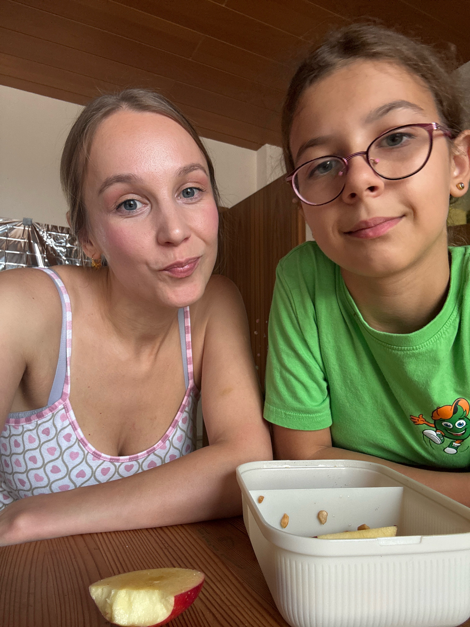 In the city of Überherrn, Germany, a young woman with blue eyes and light brown hair, wearing a white tank top with a pink and gray heart pattern, is seated at a wooden table. Beside her sits a young girl with brown hair, wearing glasses and a green t-shirt featuring a cartoon character. They are both looking at the camera. In the foreground, a slice of apple with a bite taken out of it rests on the table. To the right of the apple, a white lunchbox with dividers contains a few small pieces of food, possibly seeds and a slice of apple. The background consists of a wooden wall and a reflective surface.