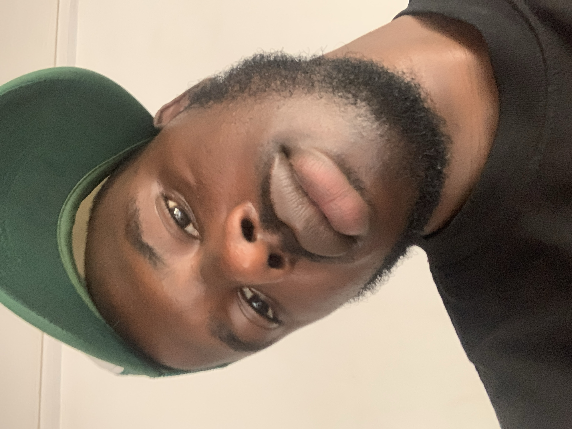 The image is a close-up portrait of a young man, captured indoors in what appears to be Jos, Nigeria, based on the provided context. His head is tilted sharply to the left (clockwise 90 degrees from an upright position), with his face dominating the frame. He has dark skin, a short, dark beard and mustache, and dark, textured hair at his hairline.
The man is wearing a dark green baseball cap, the brim and part of the crown visible on the left side of his head. He is also wearing a black t-shirt or sweatshirt, with the fabric visible around his shoulder and neck area. His features include full lips, a well-defined nose, and dark eyes that are open and directly engaging the camera. Reflections are visible in his eyes, suggesting an indoor light source.
The background is a plain, light-colored wall, providing a simple and uncluttered setting for the portrait. The man's expression is neutral to slightly contemplative, without a distinct smile or frown. There are no other visible subjects, objects, or discernible text in the image. The time of day and weather conditions are not evident due to the indoor setting.