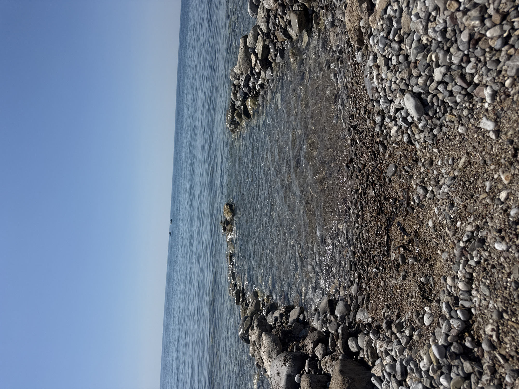 The image shows a pebble beach and a calm body of water under a clear blue sky. Two small boats are visible in the distance on the water. The foreground is filled with various sizes of pebbles and sand, with a break in the pebble line leading into the water, forming a small inlet. The water is clear, allowing the seabed of pebbles to be visible. The scene is tranquil and depicts a natural coastal environment. The location is specified as the city of Đurovići, Montenegro.