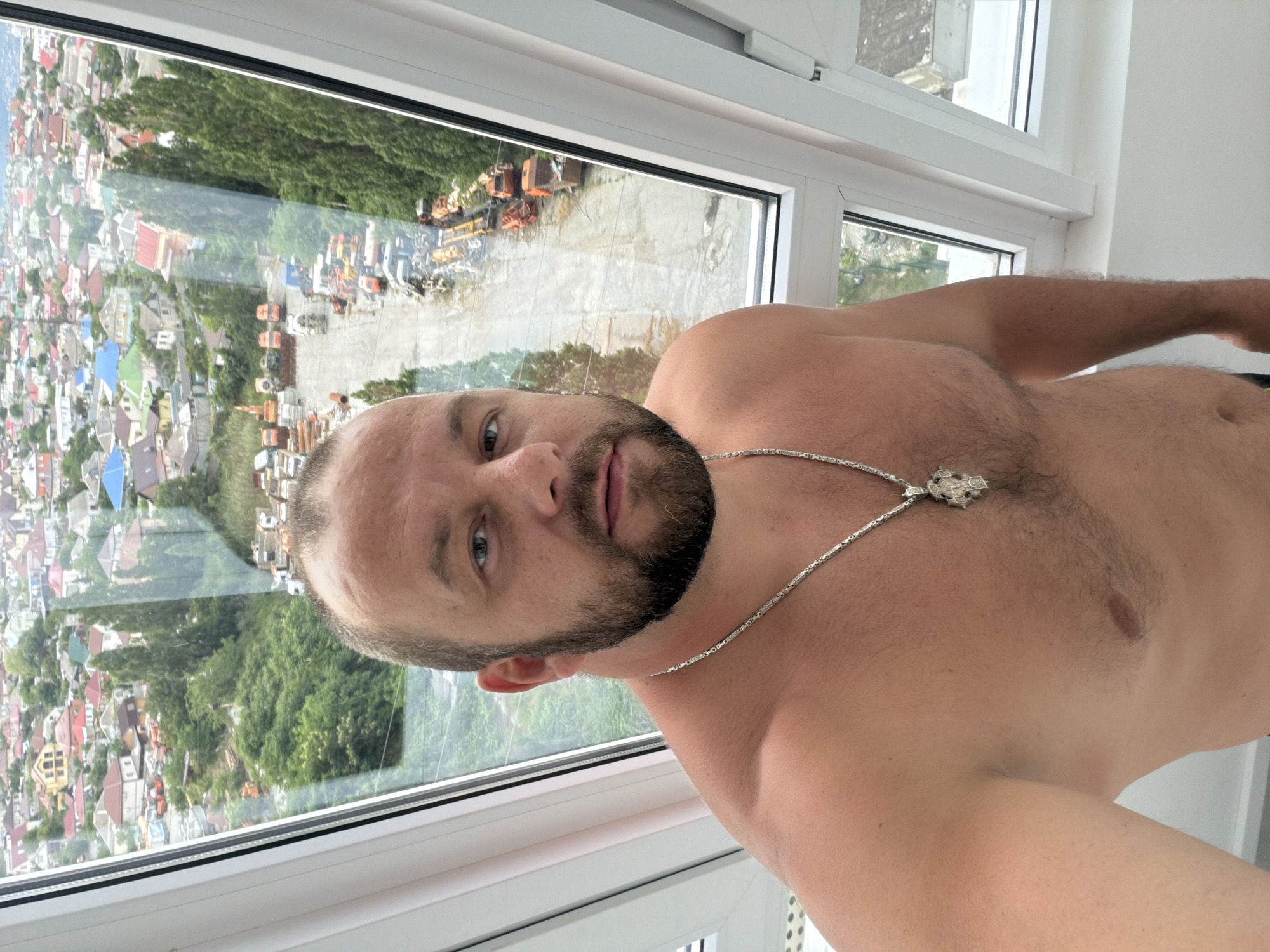 A shirtless man with a beard stands in front of a window overlooking a cityscape in Novorossiysk, Russia. The man is wearing a silver chain with a pendant. The view outside the window shows numerous houses with red and colorful roofs, green trees, and a construction site with orange vehicles and equipment. The lighting suggests it is daytime.