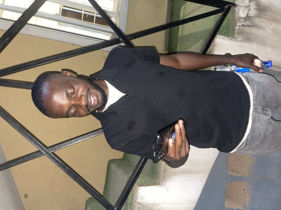 An African American man stands sideways, looking down and smiling. He holds a phone in his left hand. He is dressed in a black polo shirt with a white undershirt and gray pants. He is outside with a black metal railing behind him, and a water bottle is visible in his right hand. The background features a beige wall, some greenery, and what appears to be concrete steps.