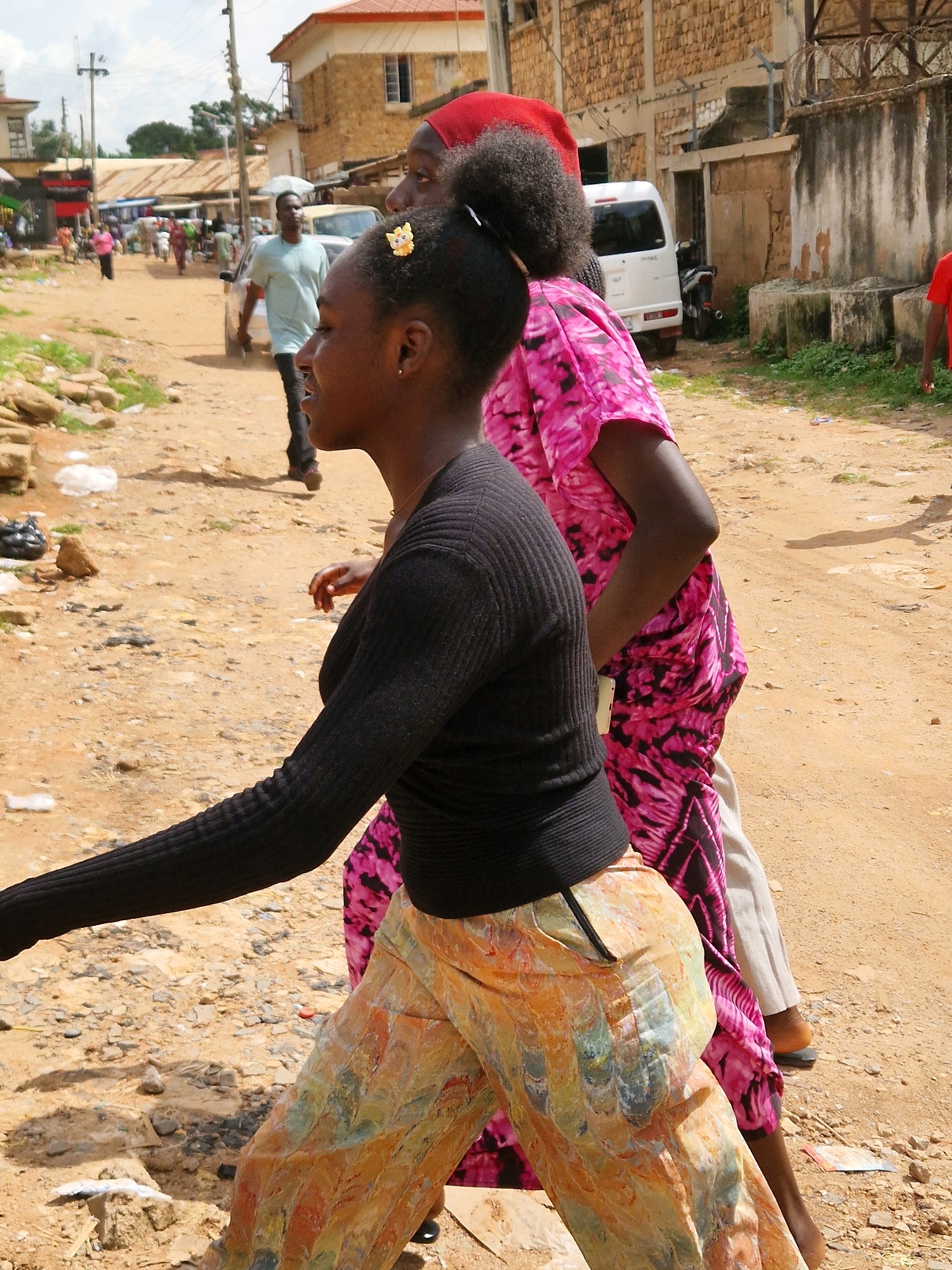 The photograph captures a street scene in Jos, Nigeria, characterized by a dirt road and the presence of several individuals. In the foreground, a young woman with dark curly hair is walking, wearing a black long-sleeved shirt and brightly colored patterned trousers. Her head is turned to her right, and she has a decorative clip in her hair. Behind her, another woman in a vibrant pink and black patterned dress is also walking.
Further in the background, a man in a light blue t-shirt and dark pants is visible, walking on the dirt road. A white van is parked on the left side of the road, and a motorcycle is parked near a building. The environment appears somewhat weathered, with loose rocks and sand on the ground, and buildings constructed from stone and concrete lining the street. The sky is overcast with scattered clouds. The overall impression is of a bustling, everyday street scene in a Nigerian urban setting.