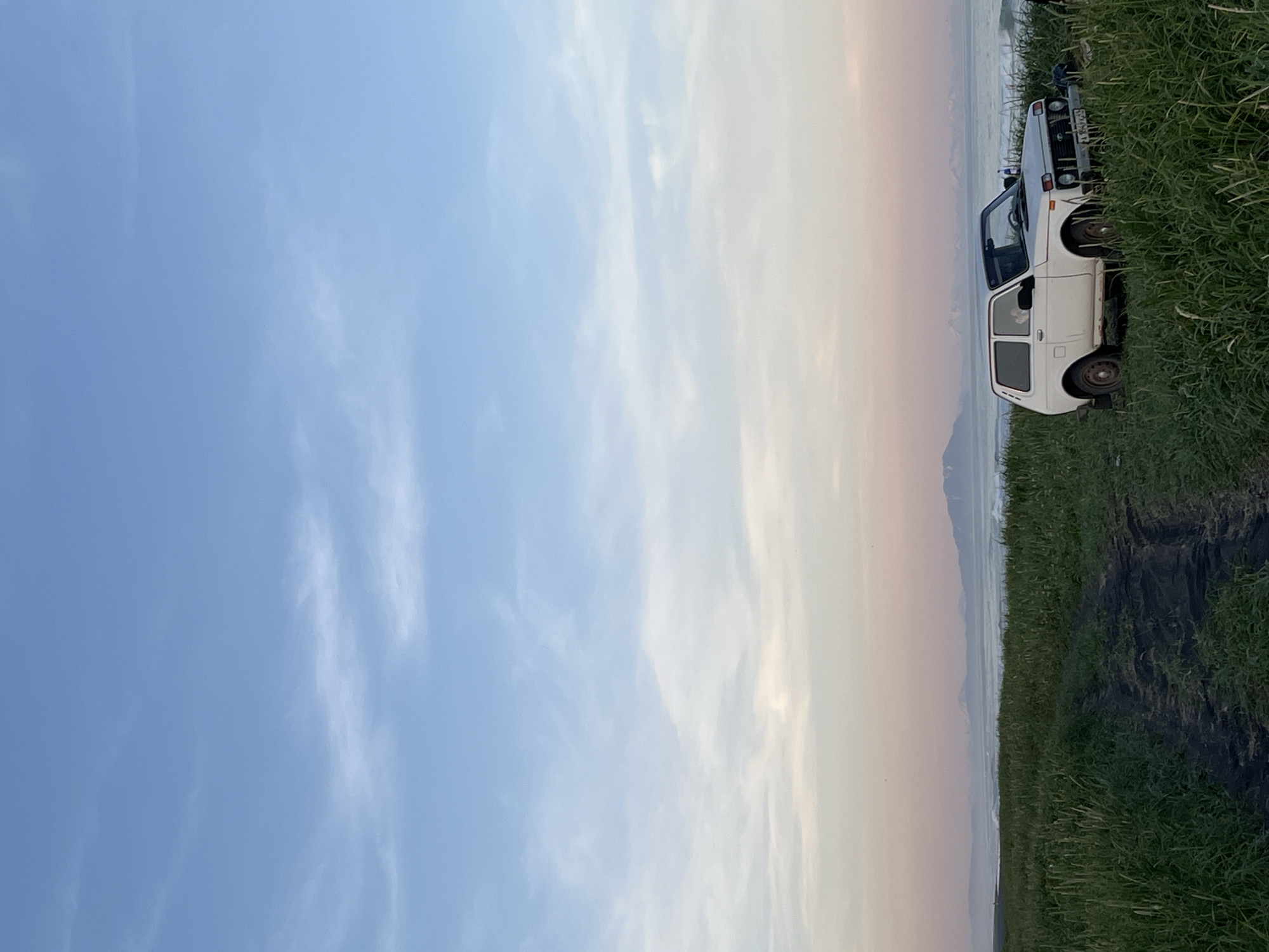 The image depicts a coastal landscape with a white SUV parked in tall green grass. The sky is a gradient of blue to pink, suggesting either sunrise or sunset. In the background, mountains are visible beyond a choppy ocean. The SUV is positioned on the right side of the frame, facing towards the ocean, with its headlights on. A dirt path leads from the foreground towards the car. The scene appears to be in a natural, undeveloped area.