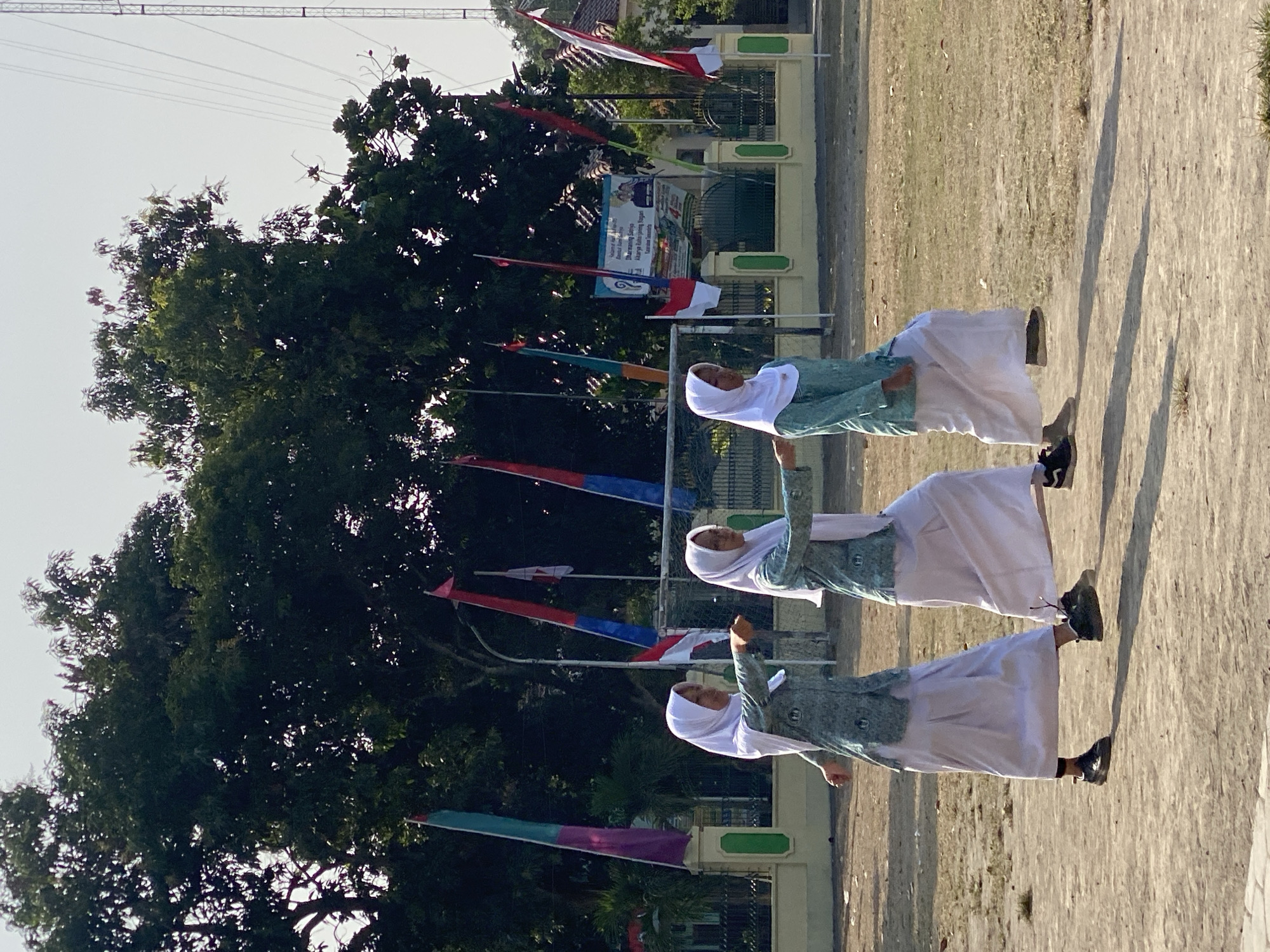 Three young females wearing white hijabs and matching teal-and-white patterned tunics over white skirts are engaged in synchronized movement, possibly a drill or dance, on a dusty outdoor field. They are positioned from left to right in the frame, with their arms extended and legs in a forward-stepping motion. The background features a low yellow wall with green trim, a tall metal tower with cables, lush green trees, and several colorful flags. One flag, on the right side, appears to be the Indonesian flag. A banner with text and images is visible on the wall. The scene suggests an event or activity taking place in a schoolyard or public space within the city of Kasihan, Indonesia. The lighting indicates it is daytime, with clear skies.
