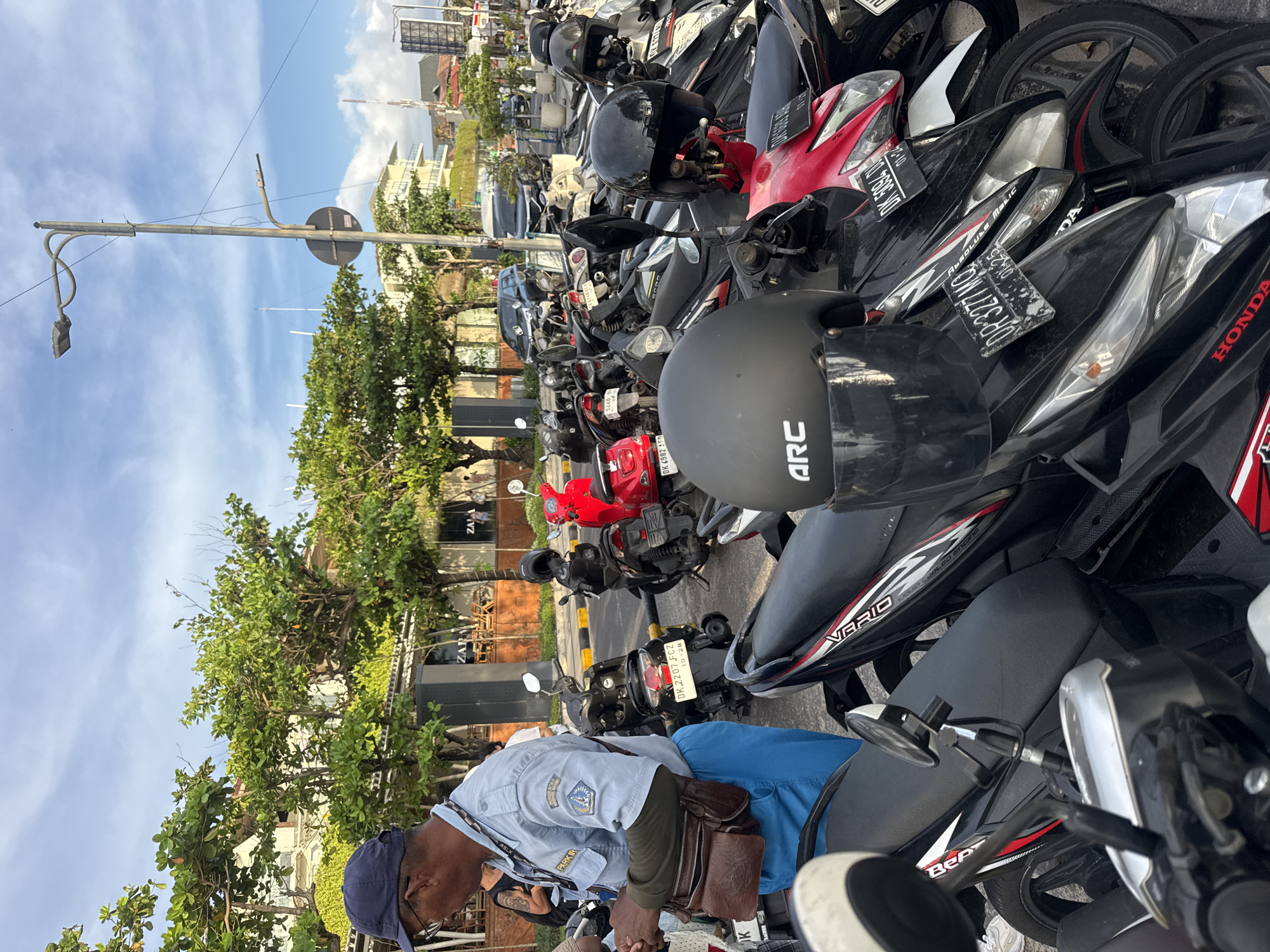 The image depicts a parking area filled with motorbikes in Kuta, Indonesia. A man in a blue shirt and cap, possibly a parking attendant, is standing amongst the parked bikes. The motorbikes are densely packed, with helmets visible on some. A sign on one of the bikes reads "DK 2207 FCZ, 10-28." "DK 4982 AFN" is visible on another bike. Brand names Honda and Vario are on others. In the background, trees and a building with "ZA" visible, indicating a Zara store, can be seen. Overhead, a utility pole stretches into a blue sky with scattered clouds.