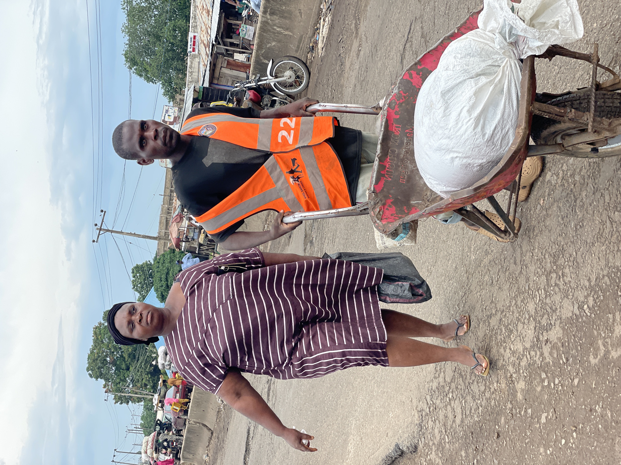 The image captures a street scene in Lokoja, Nigeria. Two individuals are the primary subjects. A man, wearing a black t-shirt and an orange reflective vest with the number "22" printed on it, pushes a wheelbarrow. The wheelbarrow is loaded with a large, white, irregularly shaped object, likely covered in plastic sheeting. The man's vest also displays a circular logo with an image of a person on it. The second subject is a woman, standing to the left of the man and slightly in front. She is wearing a dark-colored dress with vertical white stripes and a black headwrap. Her expression appears serious.
The background suggests a bustling urban environment. Shops with corrugated metal roofs line the street, and various items are visible, some on display, others as clutter on the ground. A motorcycle is parked to the left of the man, indicating common modes of transportation in the area. Power lines and poles are visible against a cloudy sky. The ground is paved with asphalt, showing signs of wear and tear.
The image appears to have been taken during the day, given the natural light. The overcast sky suggests possibly humid or rainy weather, common in tropical regions. The overall atmosphere conveys a sense of everyday activity and commerce in a busy Nigerian market or street. The woman is looking directly at the camera, while the man is looking slightly away. The image is oriented vertically, as if the photographer was holding their phone in portrait mode.
Visible text on the wheelbarrow reads "IRON CHIGO" in black capital letters, appearing reversed due to the angle.