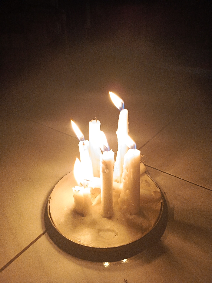 A close-up shot features a cluster of approximately eight white candles, all lit and burning brightly, serving as the main source of light in the scene. The candles vary in height and thickness, some appearing partially melted with wax dripped down their sides. They are arranged on a circular, shallow holder, which seems to be filled with melted and re-solidified wax, contained by a dark, possibly metallic, rim.
The candle holder rests on a light-colored surface, which appears to be a tiled floor with visible grout lines. The background is dark and indistinct, indicating a dimly lit environment or nighttime setting. The bright, warm glow from the flames casts soft light on the immediate surroundings, creating a warm ambiance. There are no people visible, nor any discernible text. The scene is set in Tuntang, Indonesia.