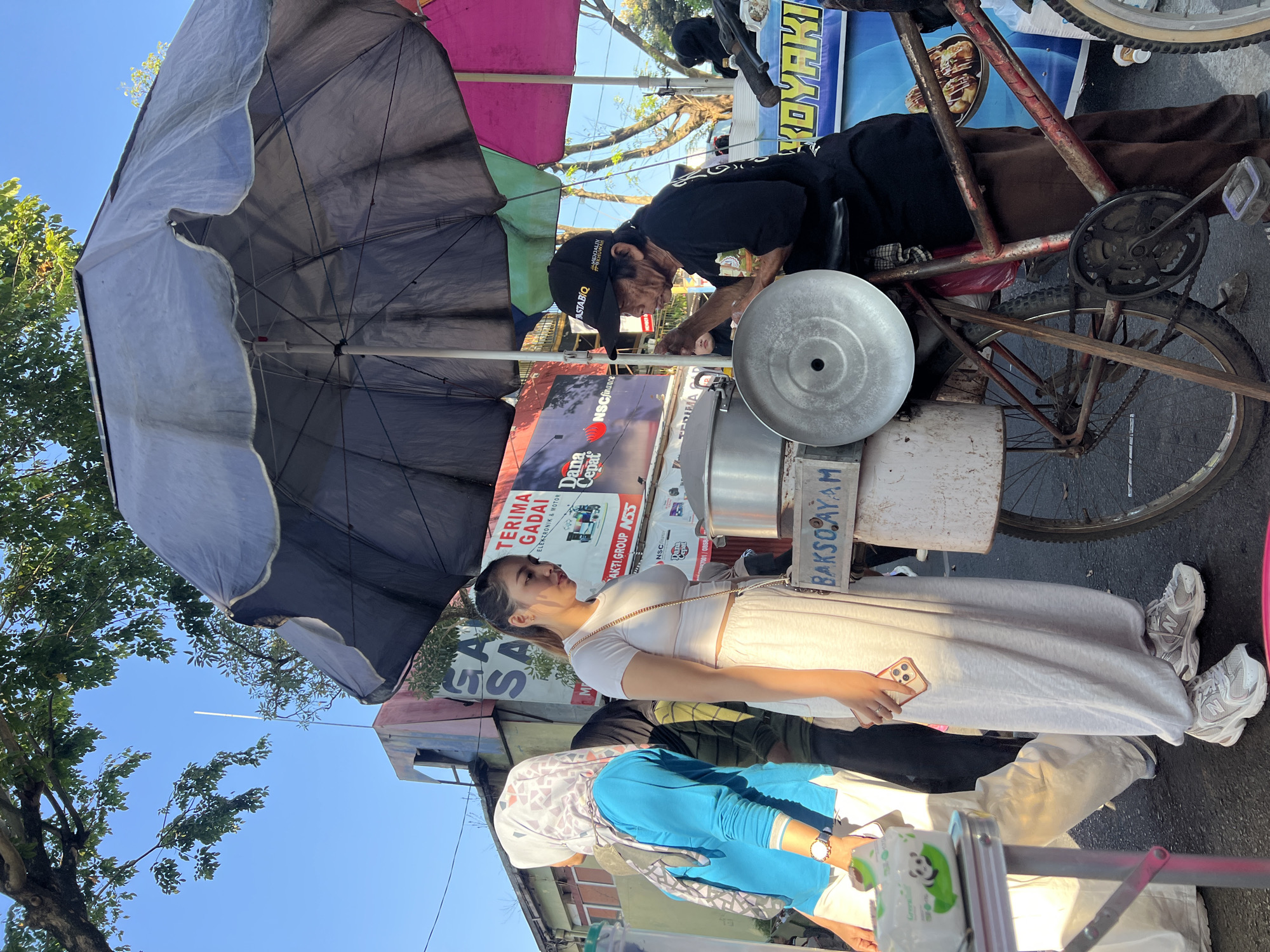 In the city of Kecamatan Pati, Indonesia, on a street, a food vendor is serving a customer. The vendor, an elderly man wearing a black t-shirt and a black baseball cap, is operating a food cart attached to a bicycle. He is holding a metal utensil and appears to be interacting with the customer. The customer, a young woman in a white crop top and white sweatpants, is holding a smartphone. She is standing in front of the food cart. Behind the customer, another woman wearing a blue top and a patterned headscarf is partially visible. The food cart has a large umbrella providing shade. Various signs are visible in the background, including "BAKSO AYAM" on the cart, and other advertisements in Indonesian. The scene is illuminated by sunlight, casting shadows on the ground. The overall environment suggests a bustling street market or roadside food stall.