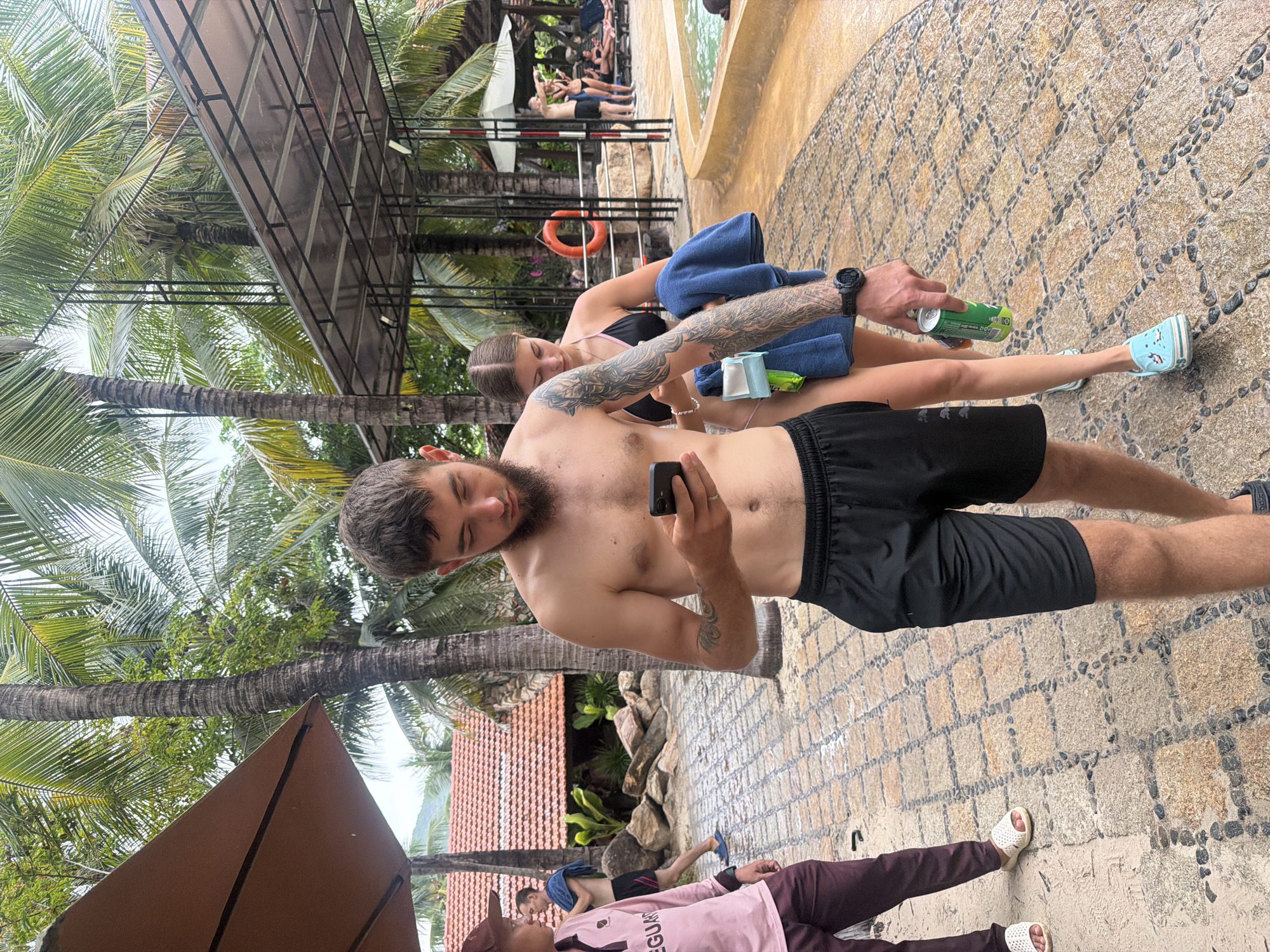 The image depicts a tropical resort environment in Nha Trang, Vietnam. In the foreground, a shirtless man with a beard and tattoos on his arm is looking at his phone. He is wearing black athletic shorts. Next to him, a woman in a black bikini is holding a blue towel and a green can. She has tattoos on her arm as well. In the background, a lifeguard in a pink shirt and burgundy pants stands near a pool area with lounge chairs and people swimming. The scene is characterized by palm trees, a tiled walkway, and the distinct red-tiled roofs of resort buildings.