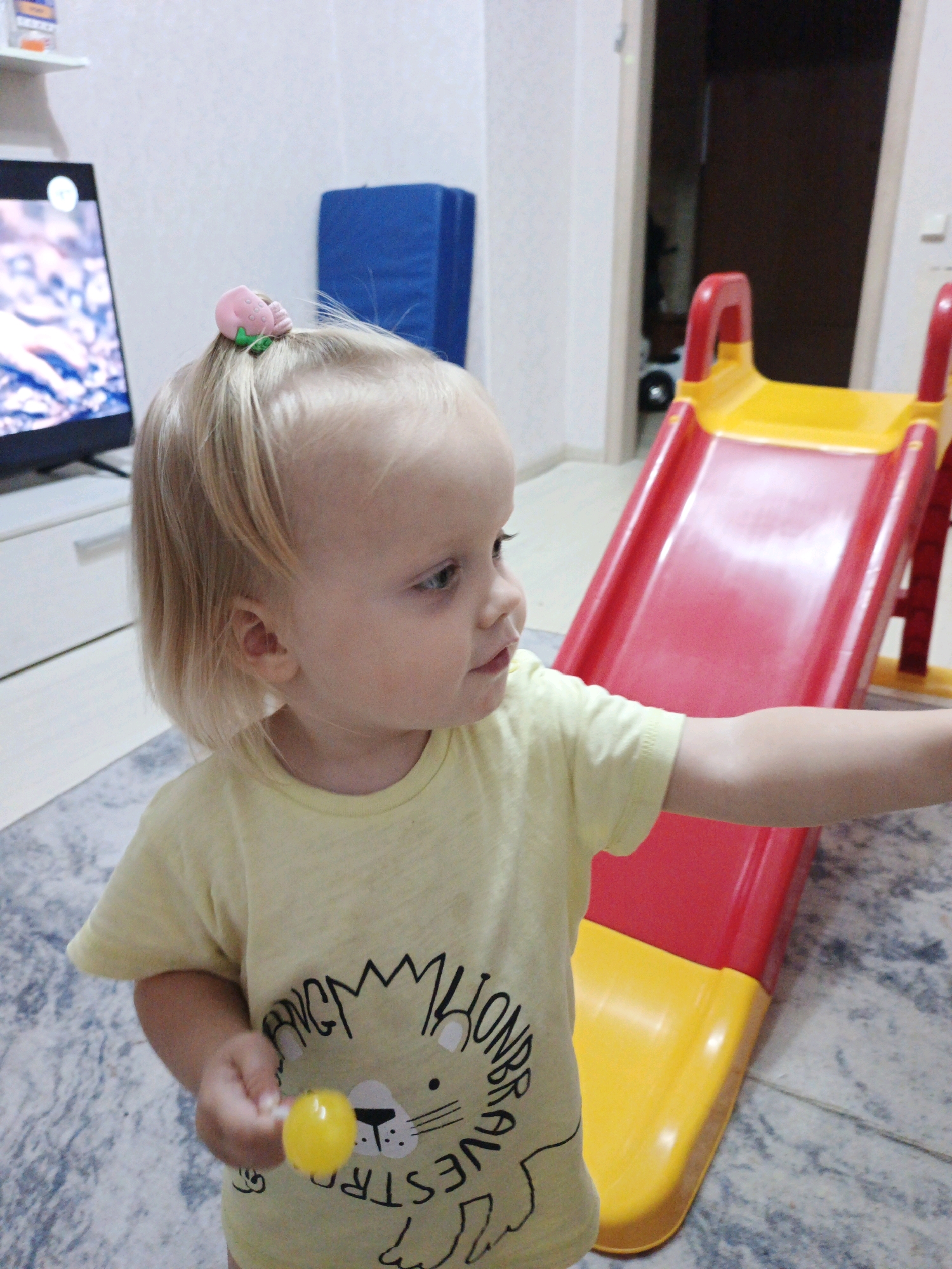 In a room, a young child with blonde hair tied with a pink strawberry-shaped clip stands in front of a red and yellow slide. The child is wearing a yellow t-shirt with a lion graphic. The child is holding a yellow lollipop in their right hand. A television screen is visible in the background, showing an image. A blue padded mat is also present in the room. The flooring appears to be tiled. The location of this scene is in the city of Zlatoust, Russia.